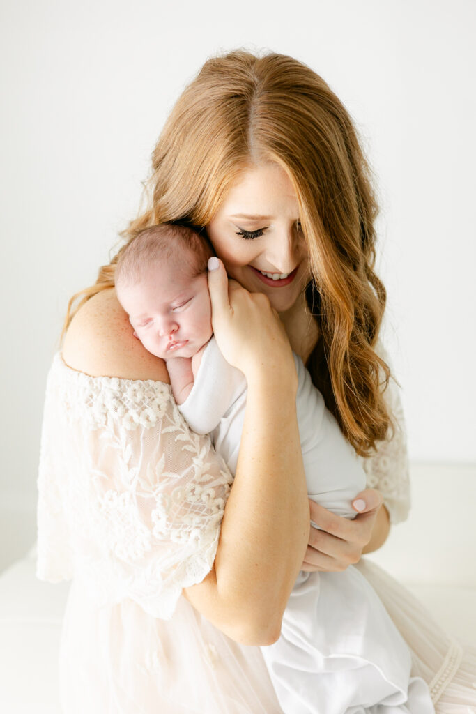 Mom is holding her newborn baby over her shoulder with her newborn wrapped in a white swaddle with mom wearing a white dress.