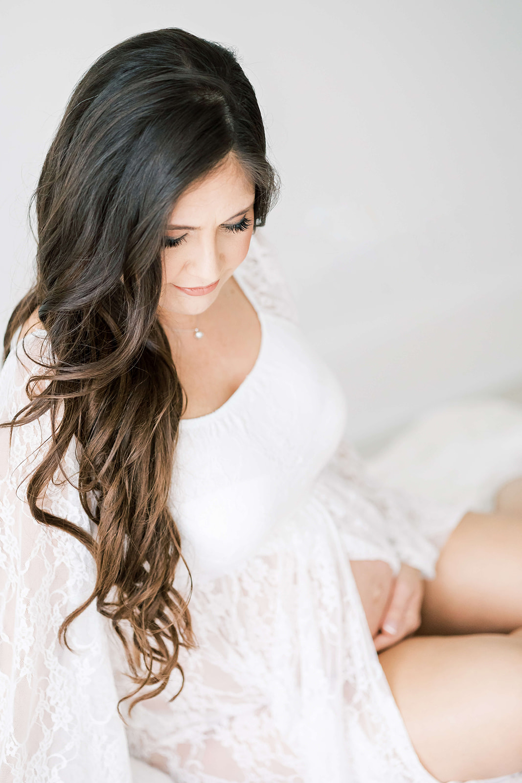 A mom to be in a white maternity gown sits on a bed in a studio