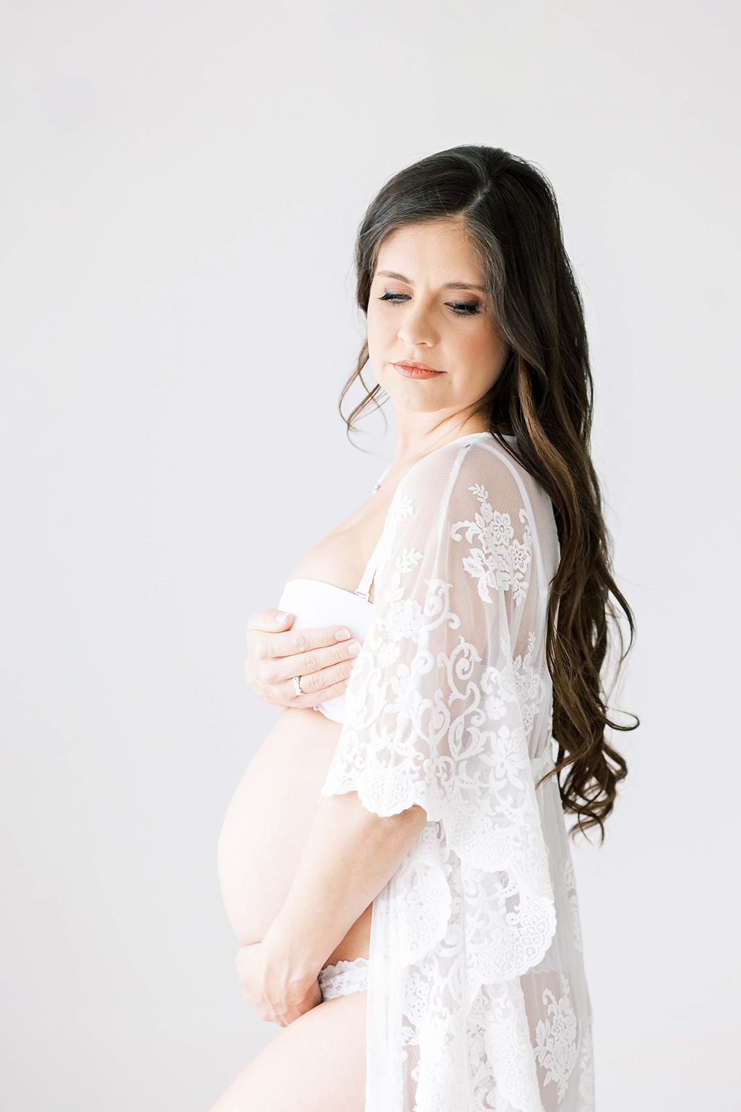 A pregnant woman in white underwear and a lace slip stands in a studio gazing down her shoulder before her 3D Ultrasound Houston