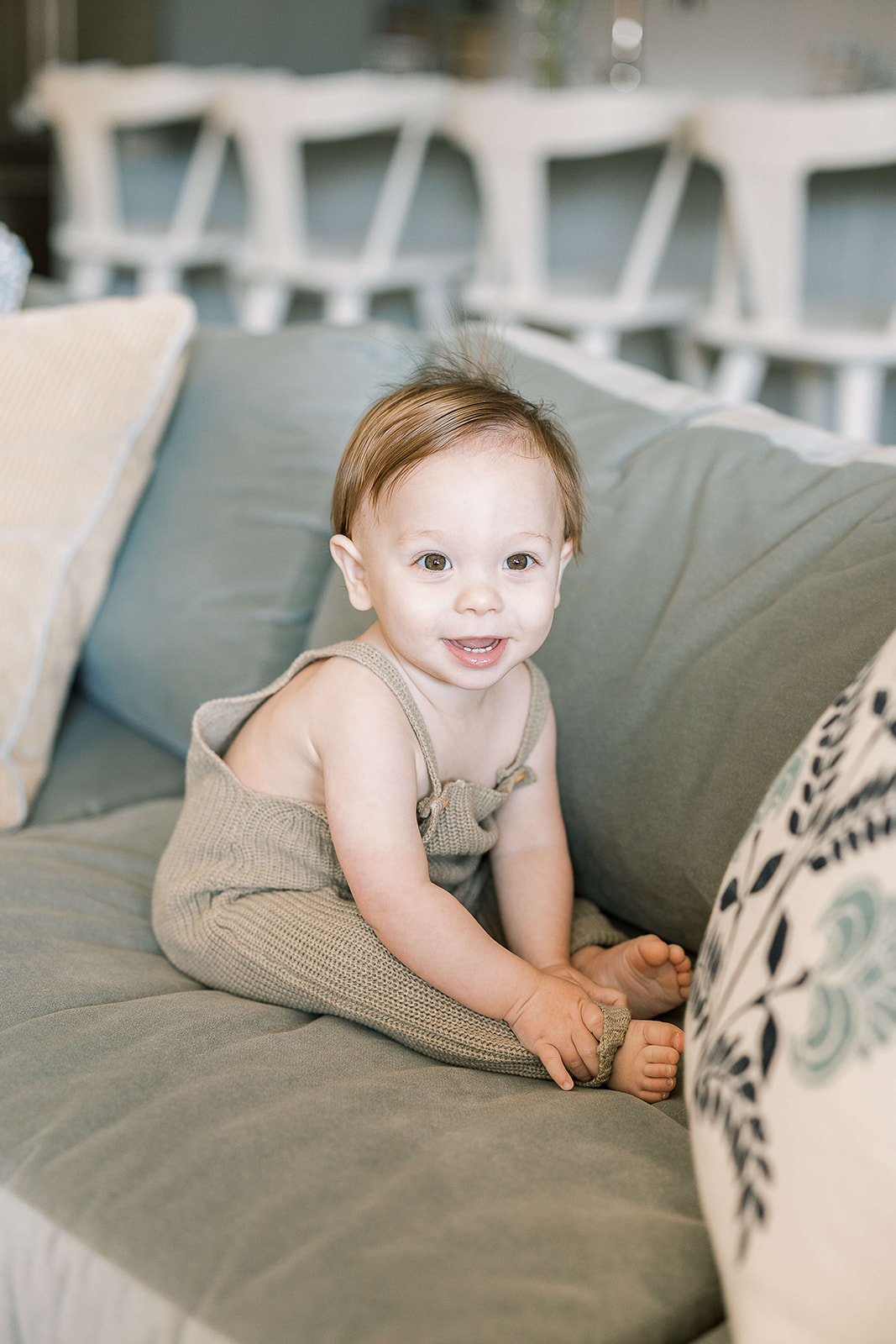 A happy infant boy in knit green overalls sits and smiles on a couch