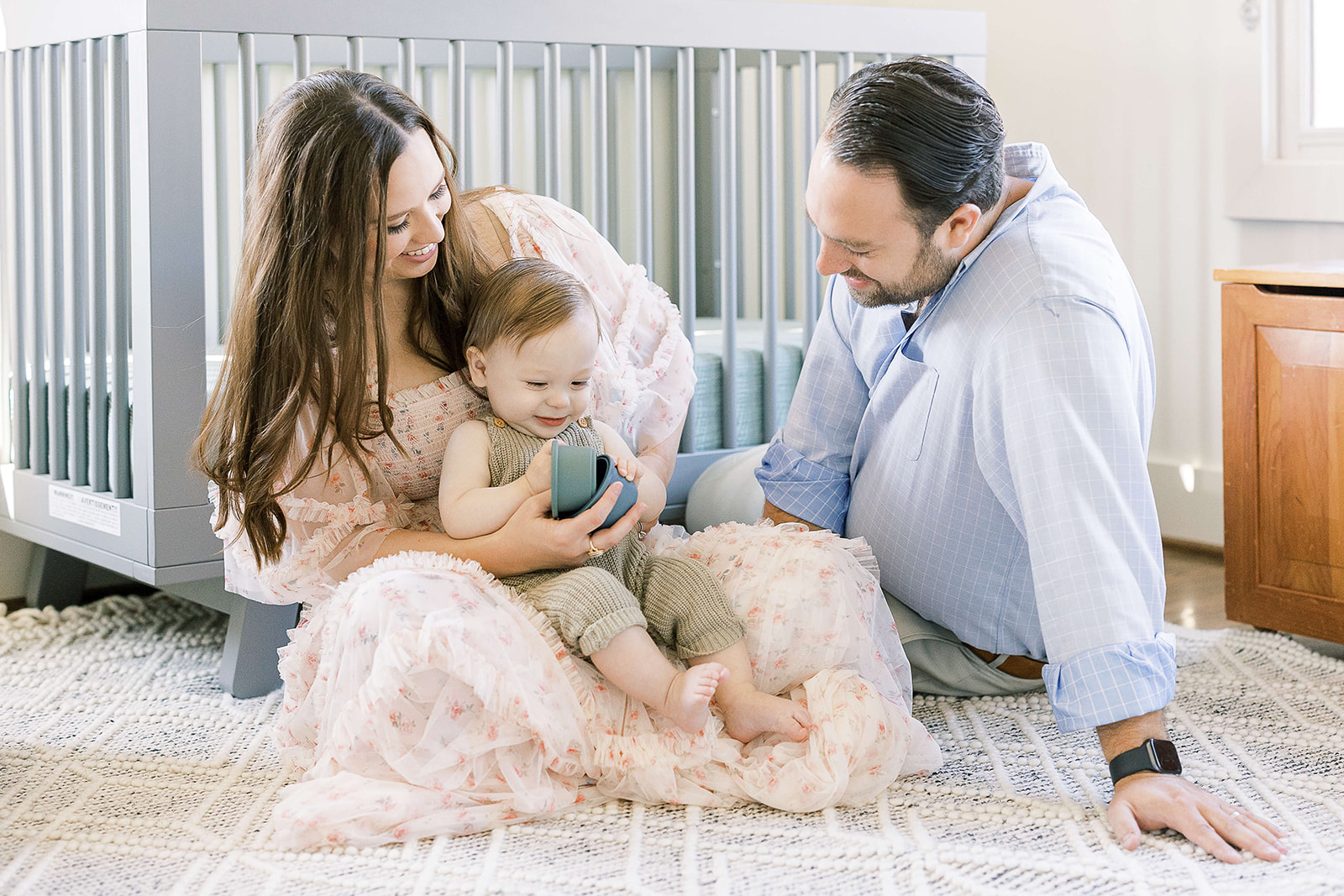 Happy mom and dad sit in a nursery playing with toys with their infant son in mom's lap after visiting Baby and Kids 1st