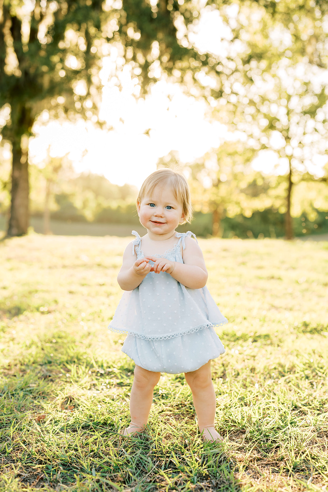 An infant girl in a blue dress stands in a grassy park at sunset after some Baby Music Classes Houston