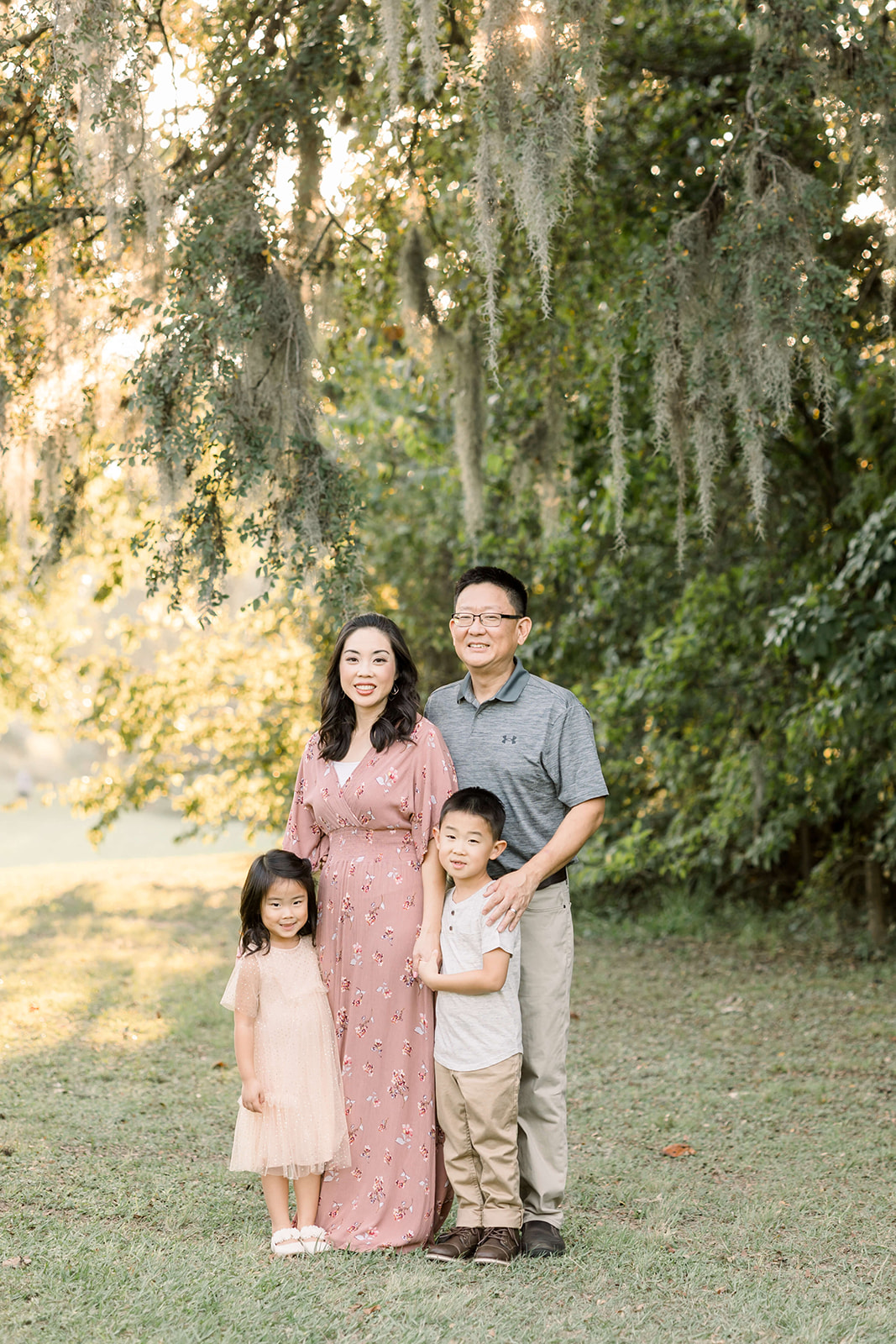 Happy mom and dad stand in a park at sunset with their toddler son and daughter under an oak tree