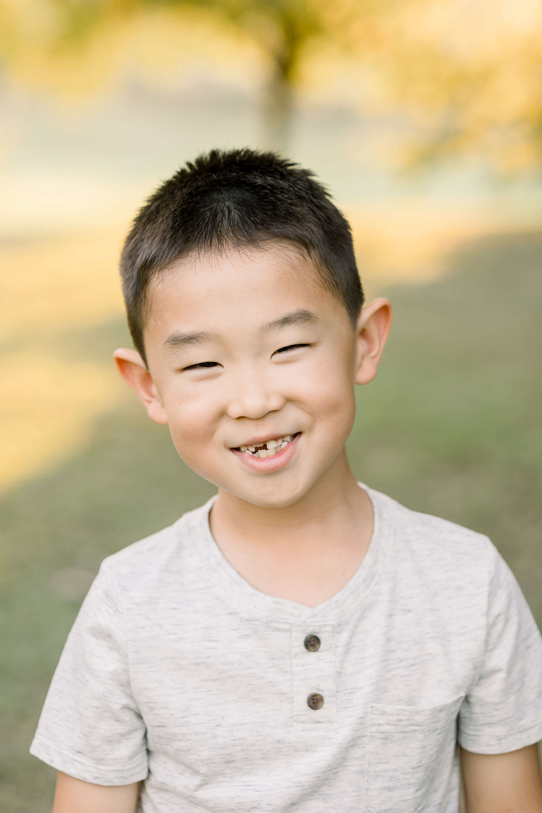 A toddler boy smiles big while missing teeth in a grey shirt in a park at sunset
