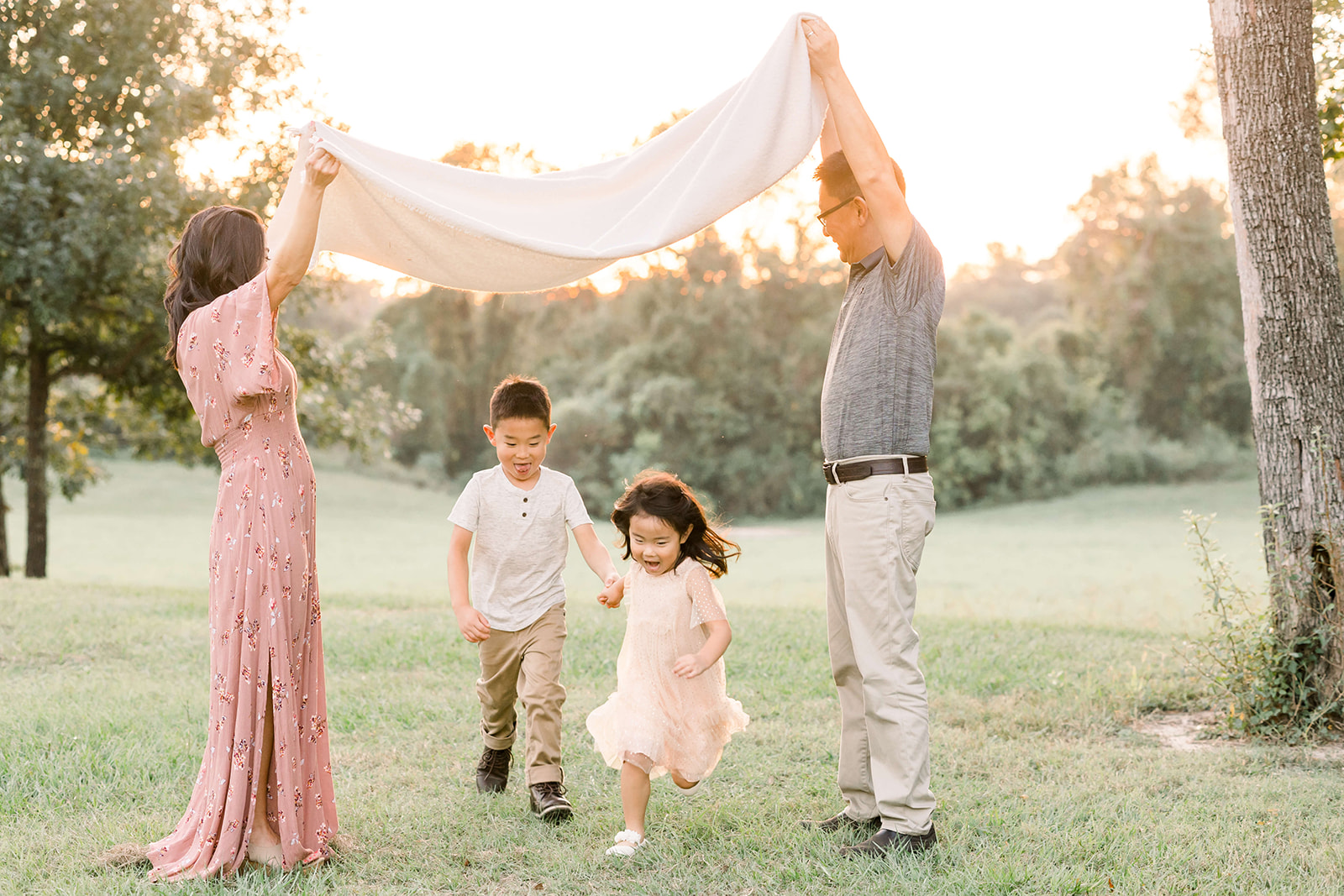 A toddler boy and his younger sister run under a blanket holding hands being held up by mom and dad in a park at sunset after visiting Dentini Pediatric Dentistry