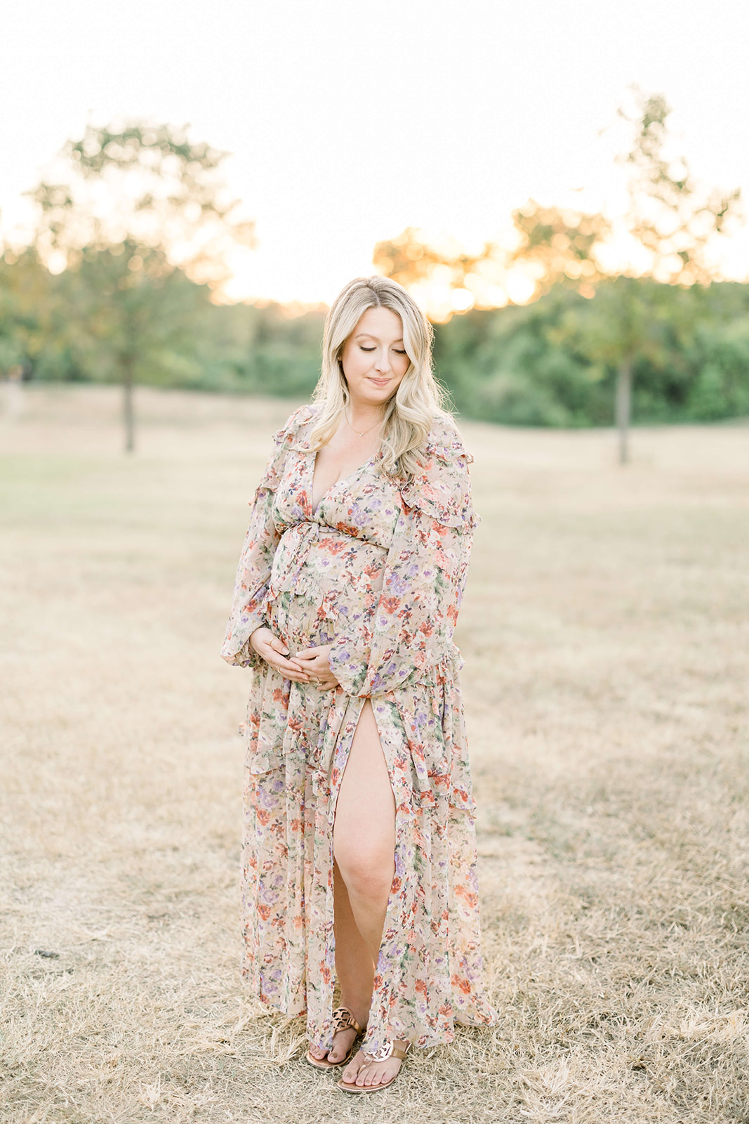 A mother to be smiles down her shoulder while walking through a park at sunset in a floral print dress