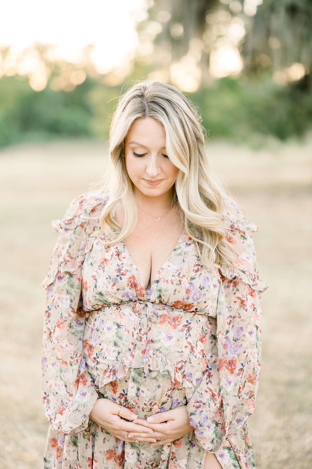 A mother to be in a floral print dress stands in park at sunset smiling down at her bump in her hands after visiting a Houston Birthing Center