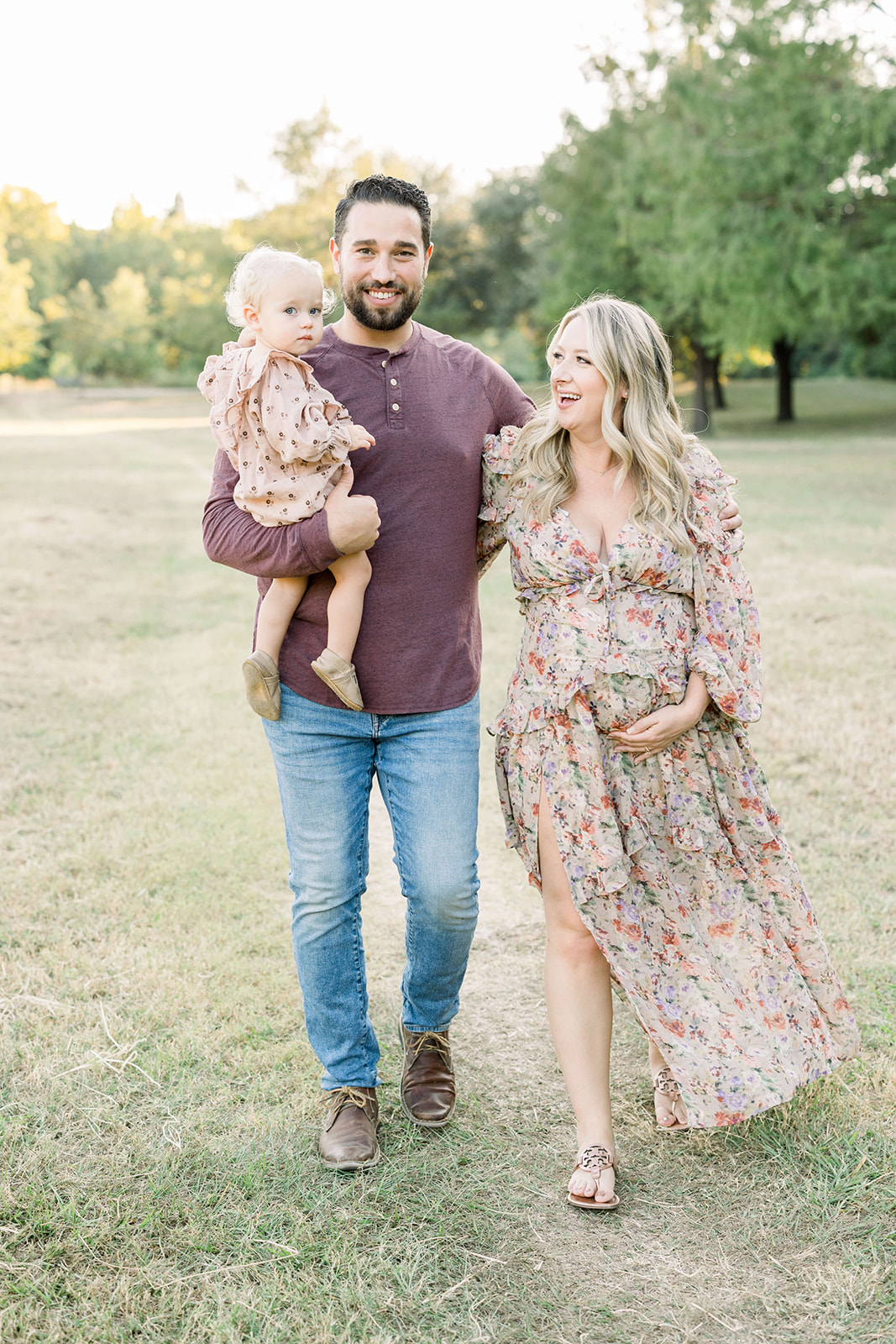 Happy expecting parents walk arm in arm through a park with their toddler daughter in dad's arm and mom holding bump after visiting a Houston Birthing Center