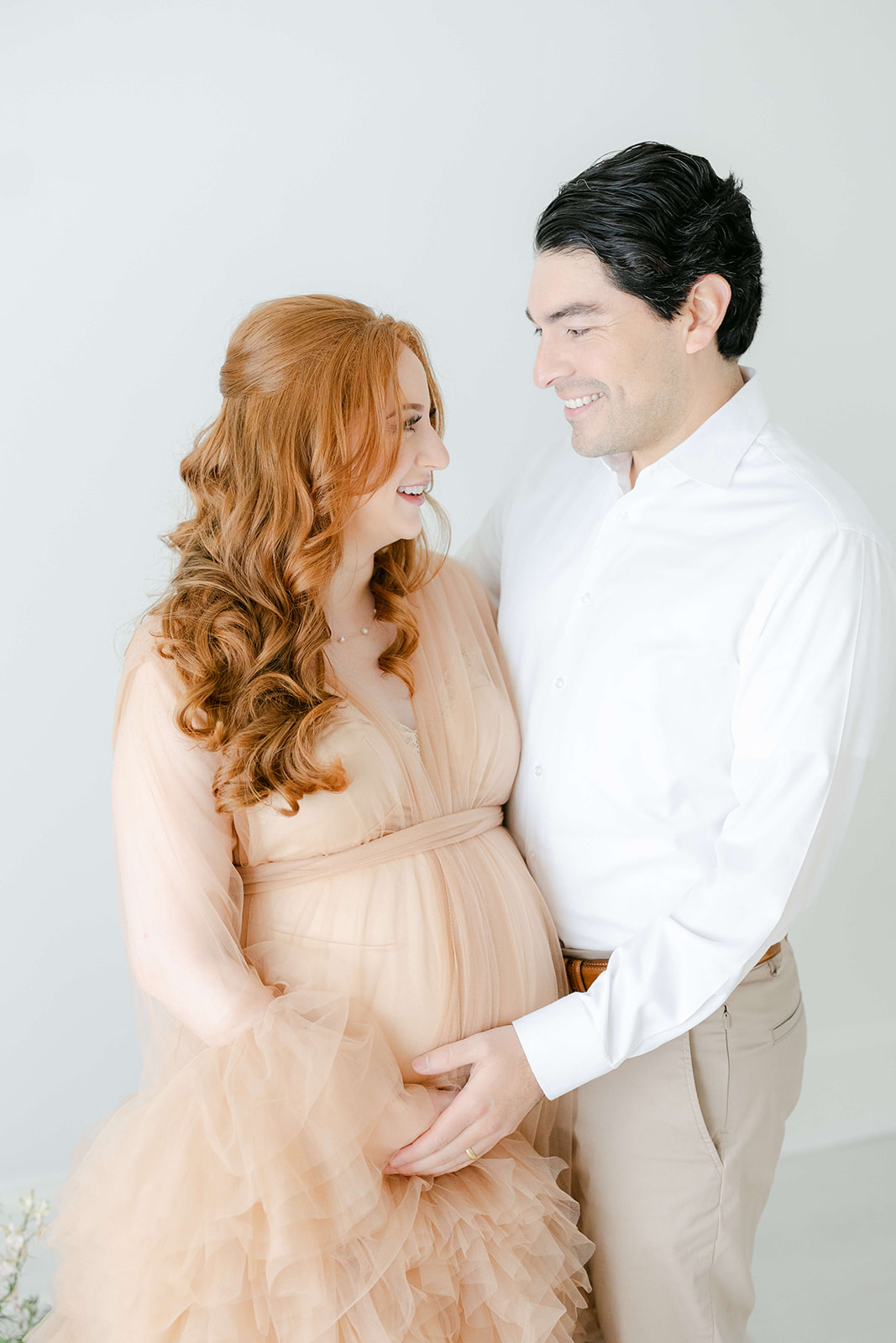 A mother and father to be stand in a studio holding the bump and smiling at each other