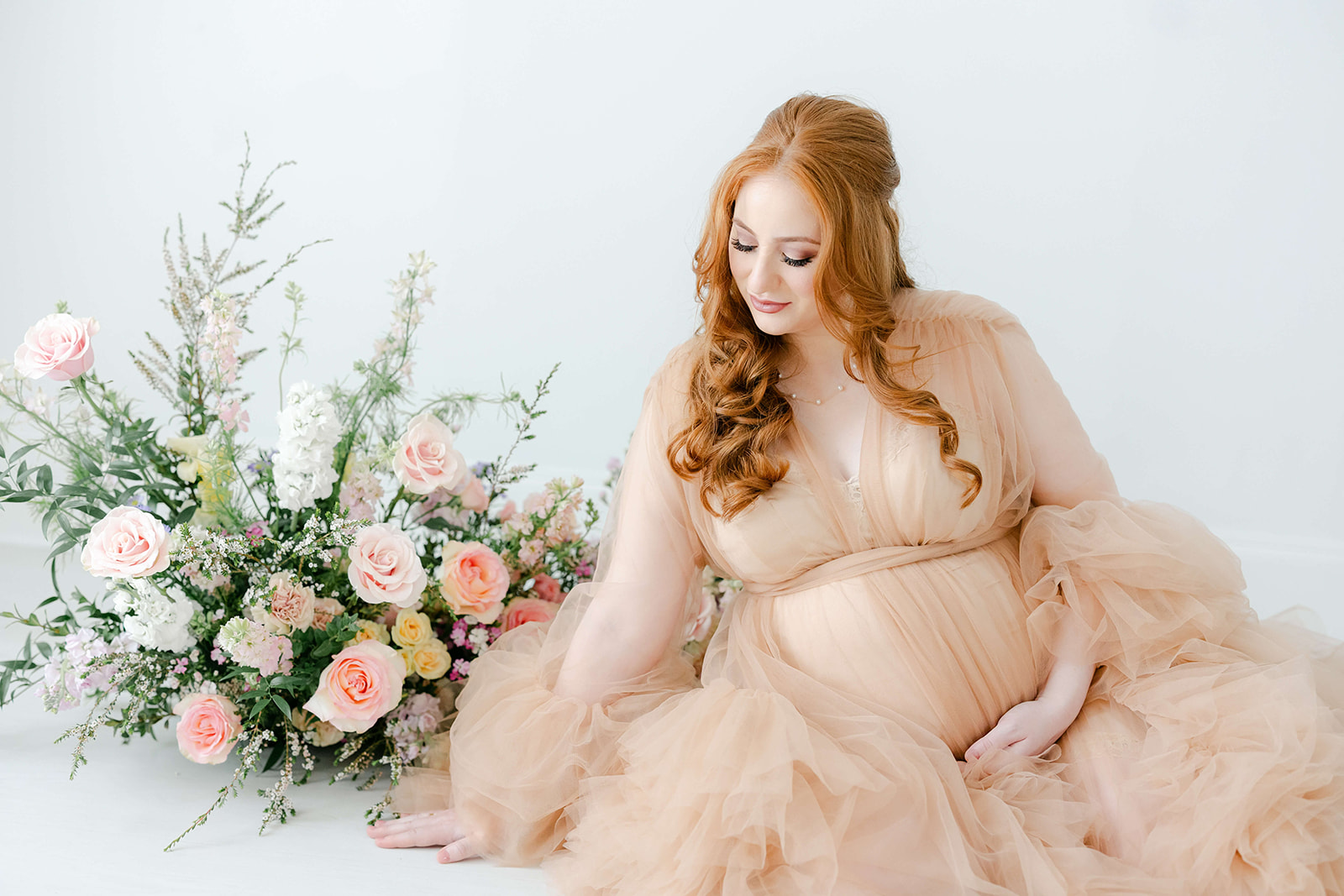 A mother to be sits on the floor of a studio with a pink bouquet of flowers with long red hair in a pink maternity gown