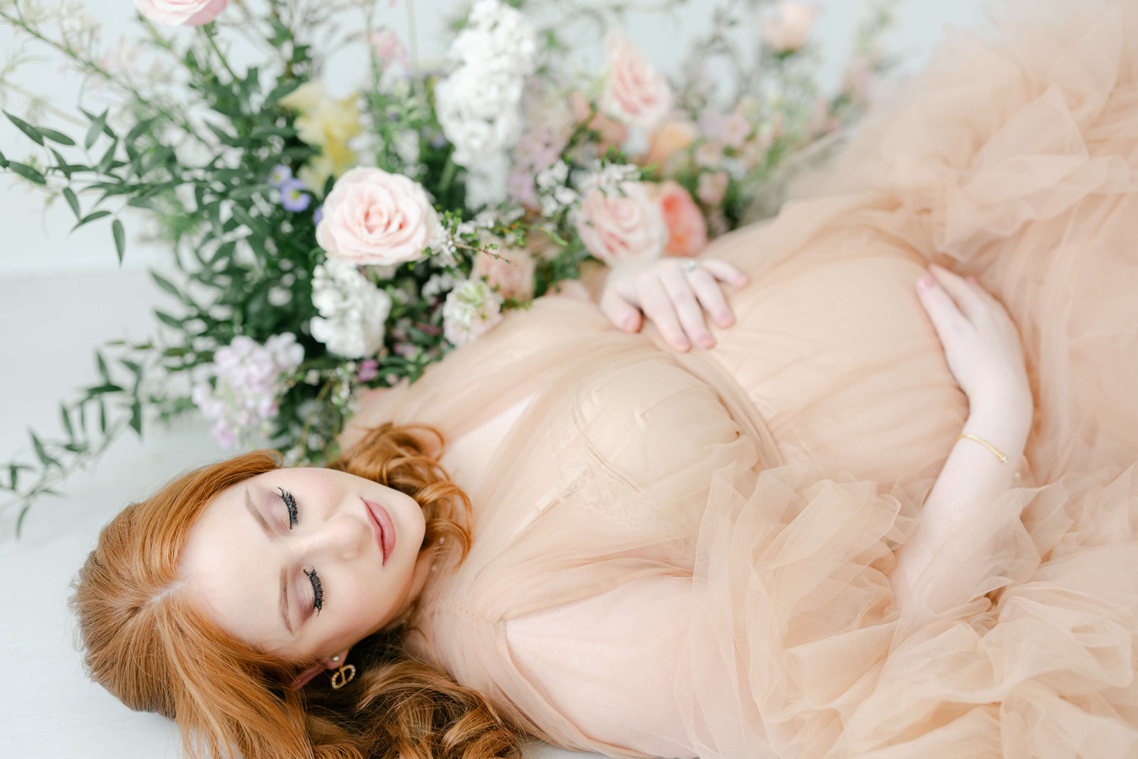 A mom to be smiles down to her bump in her hands while wearing a pink maternity gown with a large bouquet of flowers after meeting Houston Doulas
