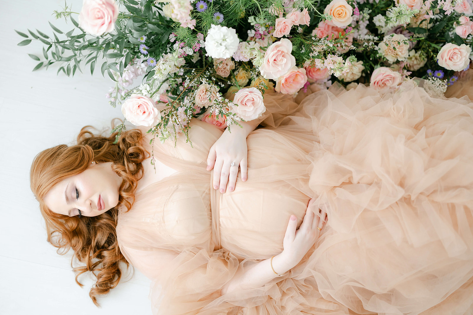 A mother to be with red hair lays on the floor of a studio surrounded by flowers in a pink tule maternity gown after visiting with Houston Doulas