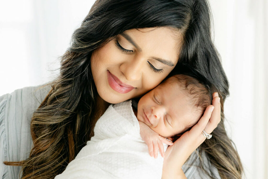 A happy new mom snuggles her smiling and sleeping newborn baby in a studio