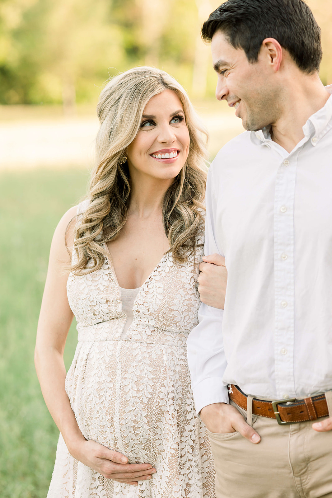 A mom to be holds her bump while holding onto her husband's arm and smiling up to him in a park at sunset