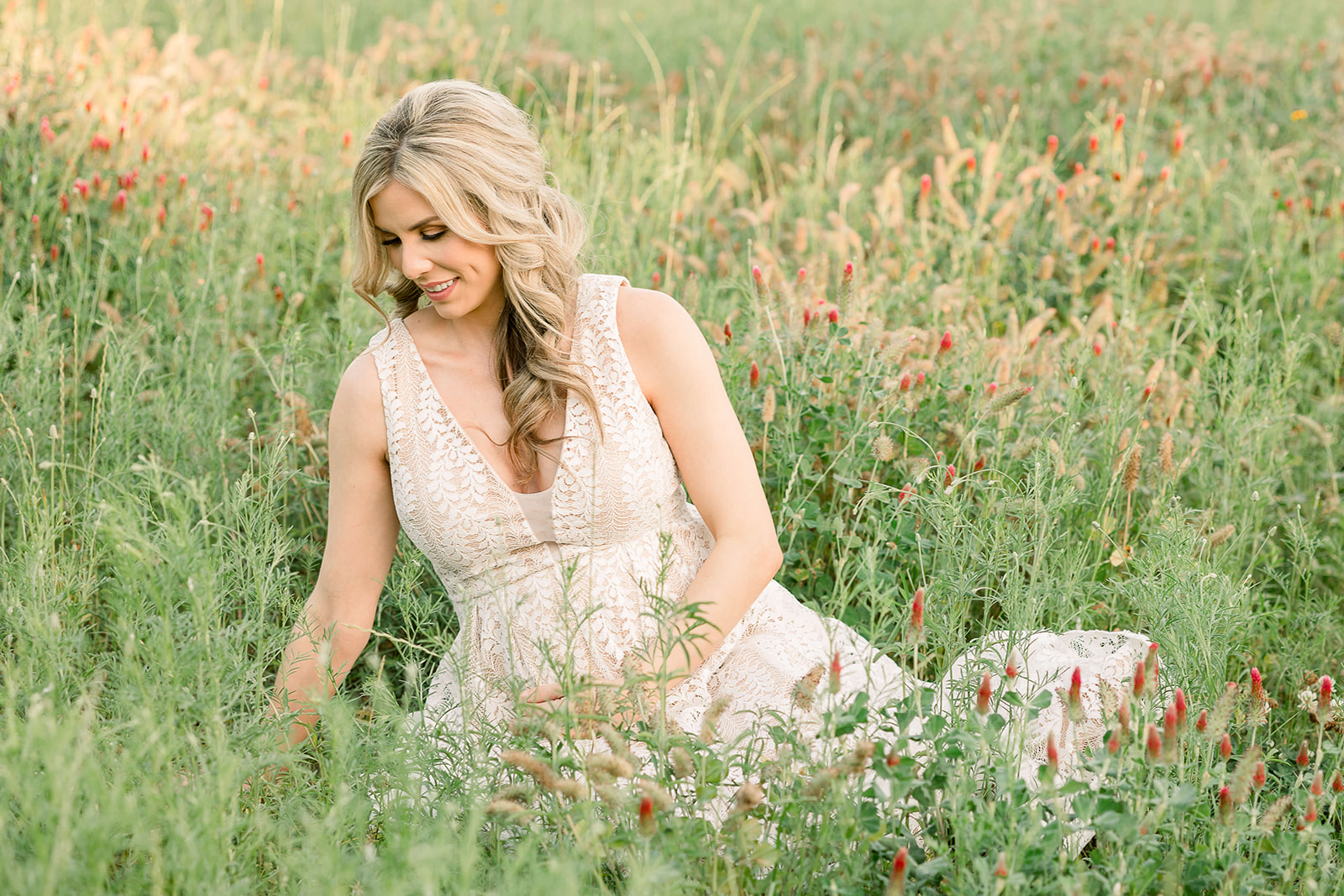 A mom to be in a beige lace maternity gown sits in a field of red wildflowers while smiling