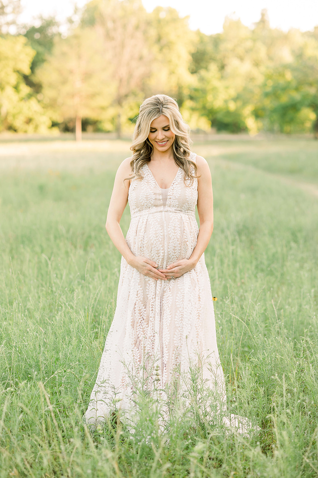 A happy mom to be in a lace maternity gown smiles down at her bump while standing in a field of tall grasses after meeting Houston Midwives