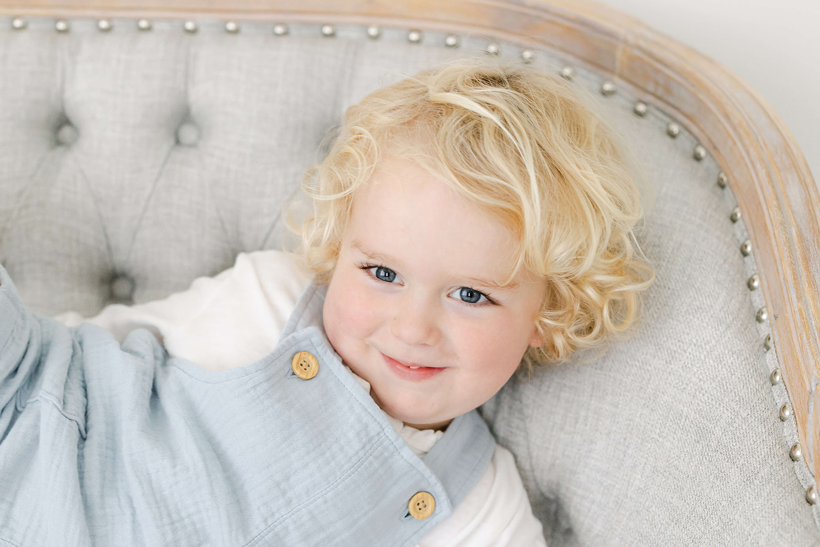 A toddler with long blonde hair plays on a bench in a studio in blue overalls