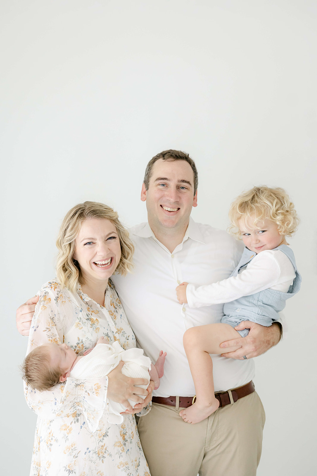 A father stands laughing in a studio while holding his toddler on his hip and mom holding their newborn baby in his other arm after meeting Houston Nannies