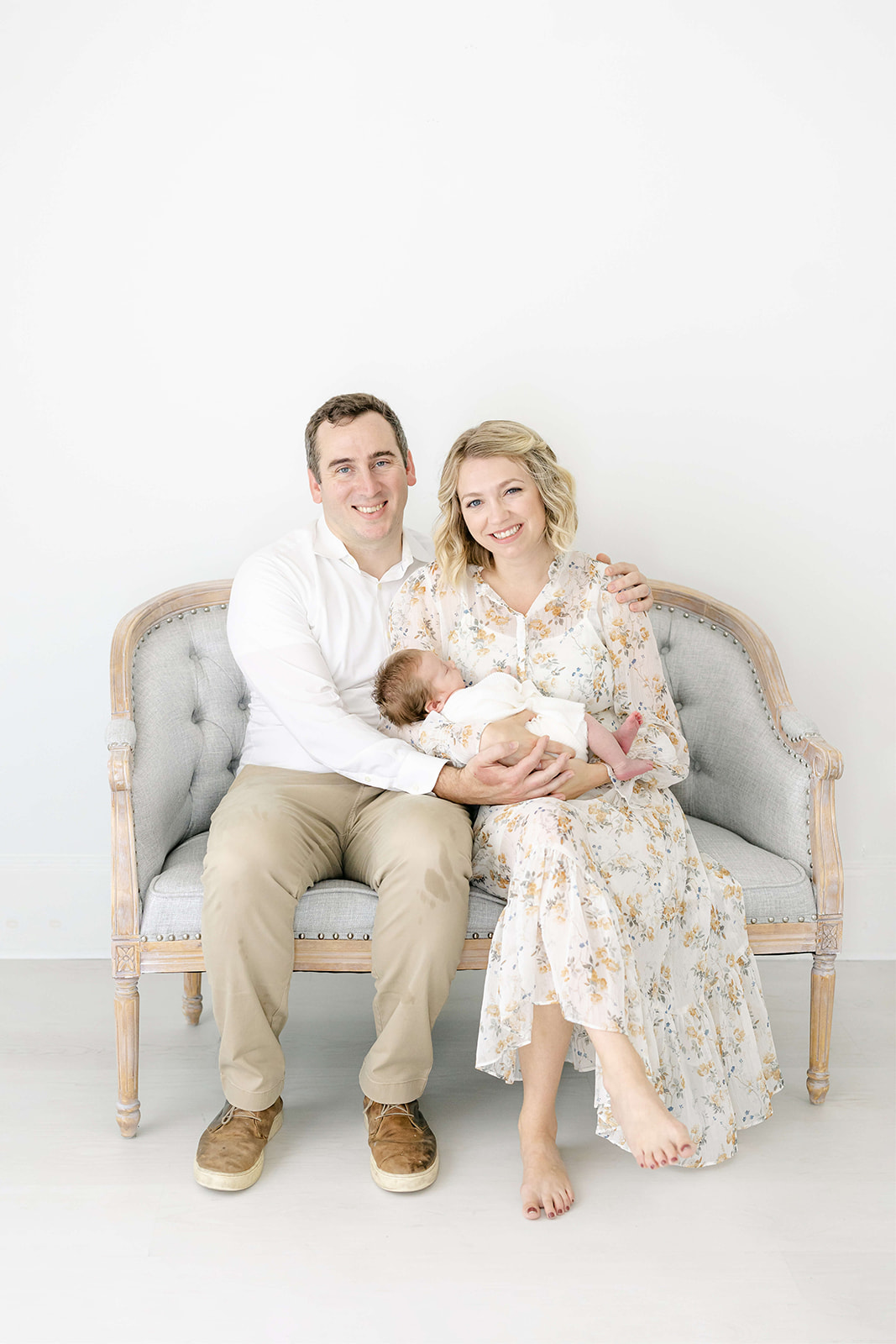 Happy parents sit on a bench in a studio holding their sleeping newborn baby and smiling after finding Houston Nannies