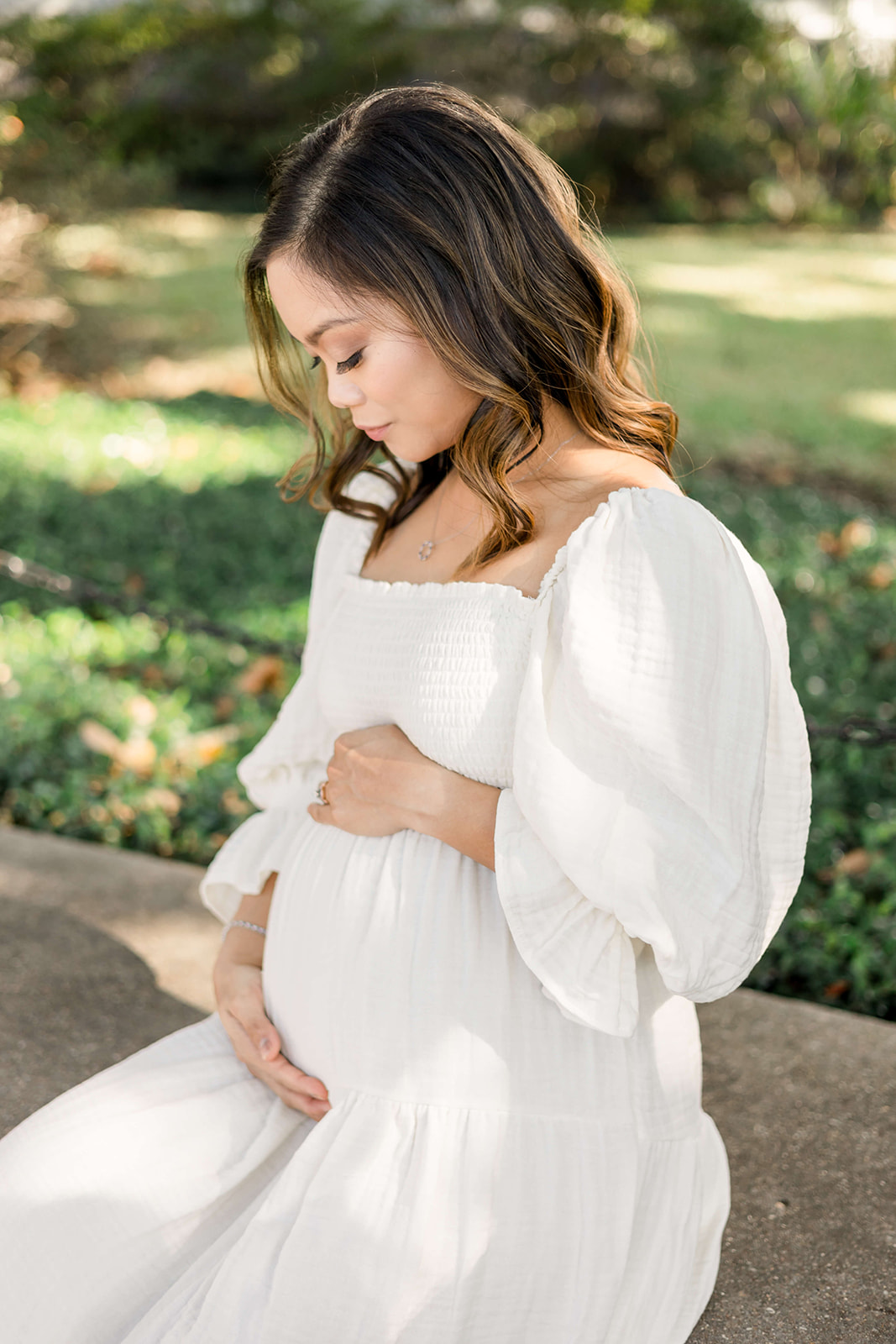 A happy mom to be sits on a garden bench holding her bump in a white maternity gown