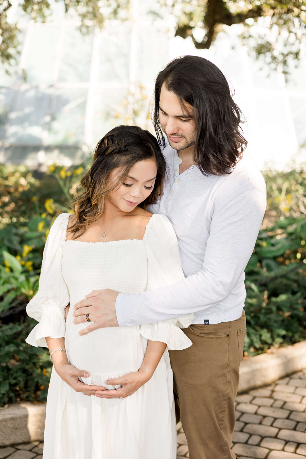 Happy expecting parents snuggle while standing in white in a garden after visiting North Houston Birth Center