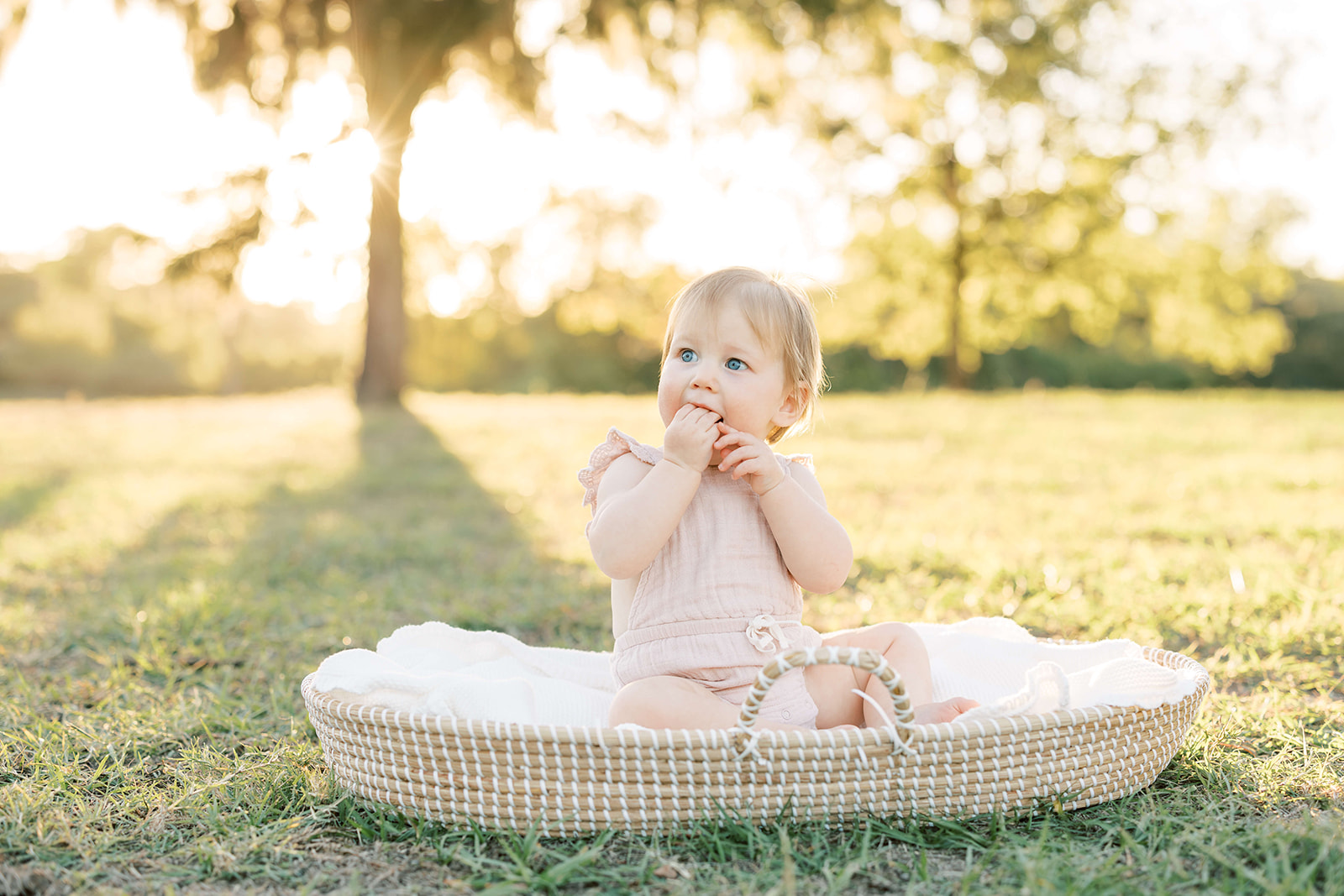 A toddler girl in a pink onesie sits in a woven basket chewing her hand at sunset in a park