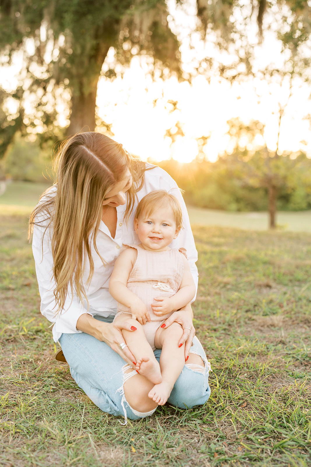 A happy mom kneels in a park at sunset with her happy toddler daughter in her lap after visiting The Itsy Bitsy Boutique