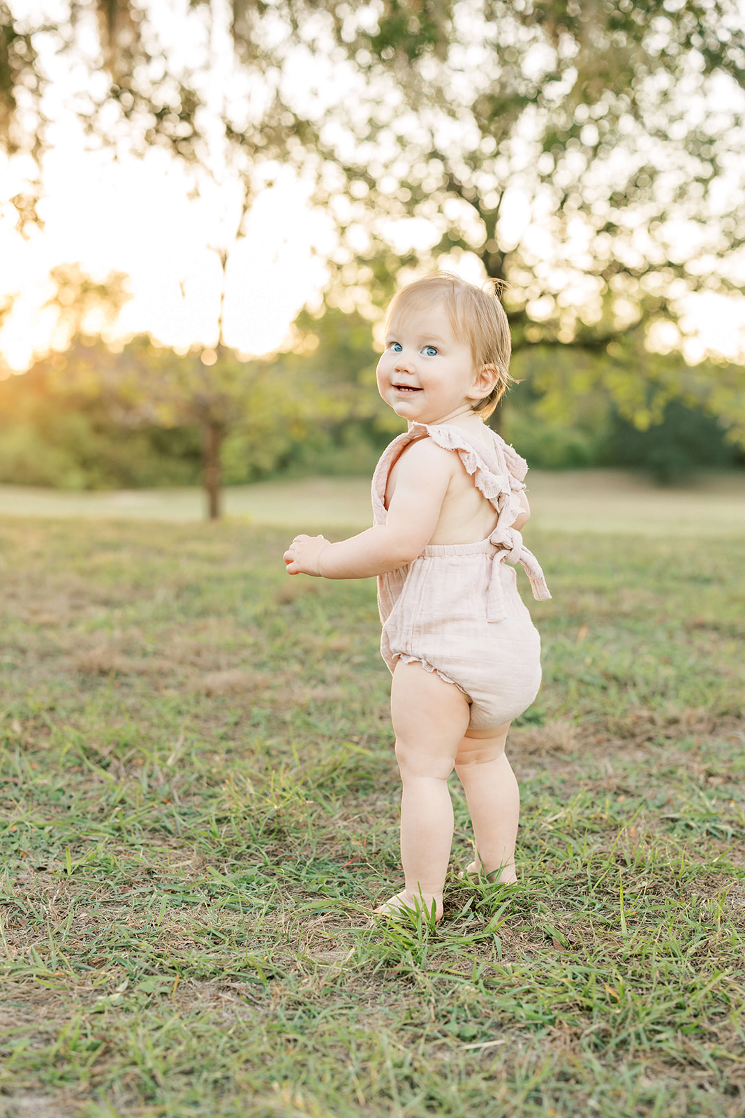 A toddler girl smiles over her shoulder while exploring a park at sunset in a pink onesie after visiting The Itsy Bitsy Boutique