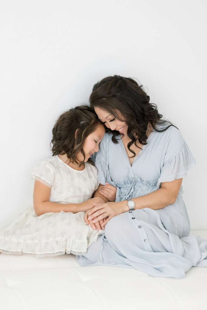 A mother in a blue dress snuggles with her toddler daughter in a white dress on a bed in a studio