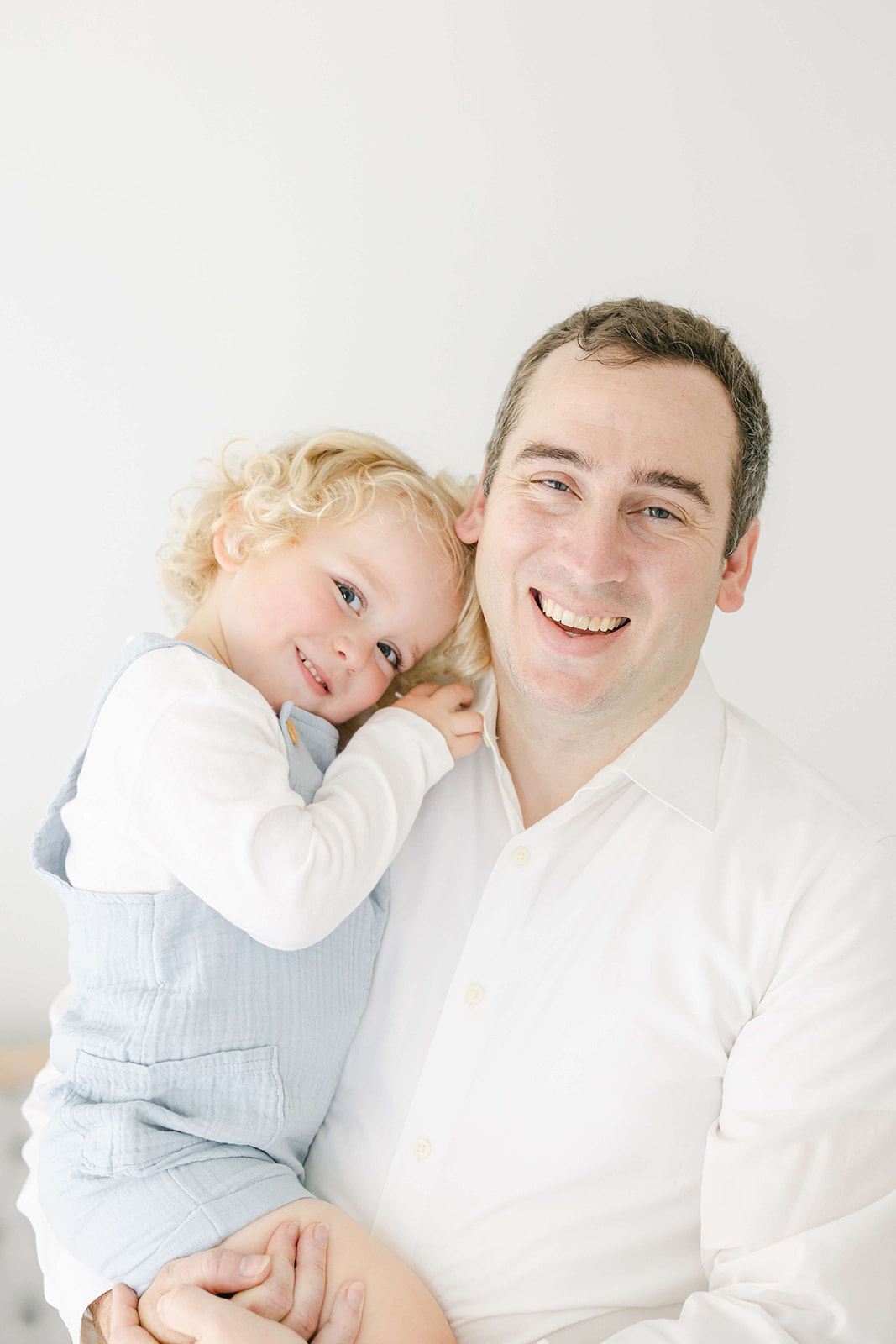 A toddler hugs onto daddy as he laughs and stands in a studio