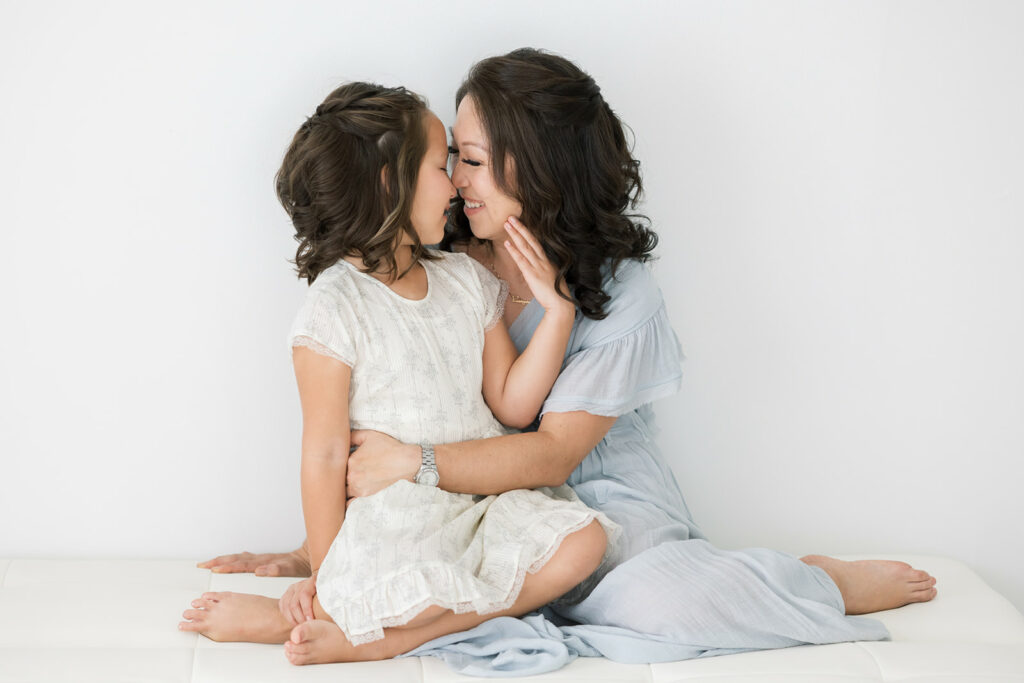 A mother and daughter snuggle while sitting on a white bed in a studio after shopping at Threadfare