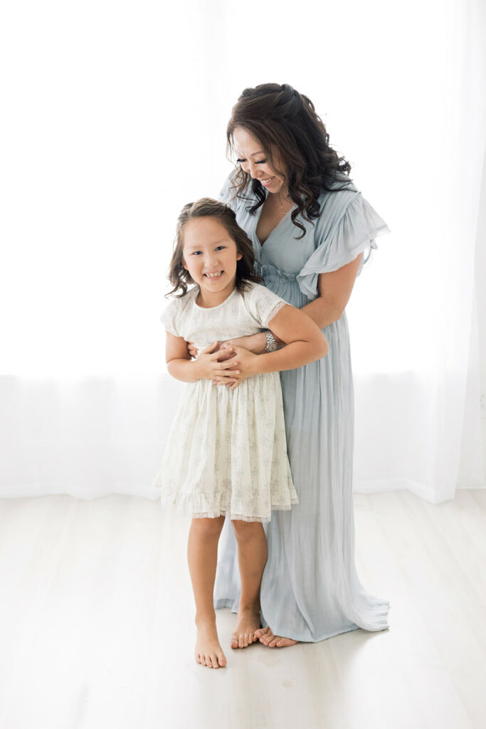 A mom and daughter both in dresses from Threadfare laugh and play in a studio in front of a window
