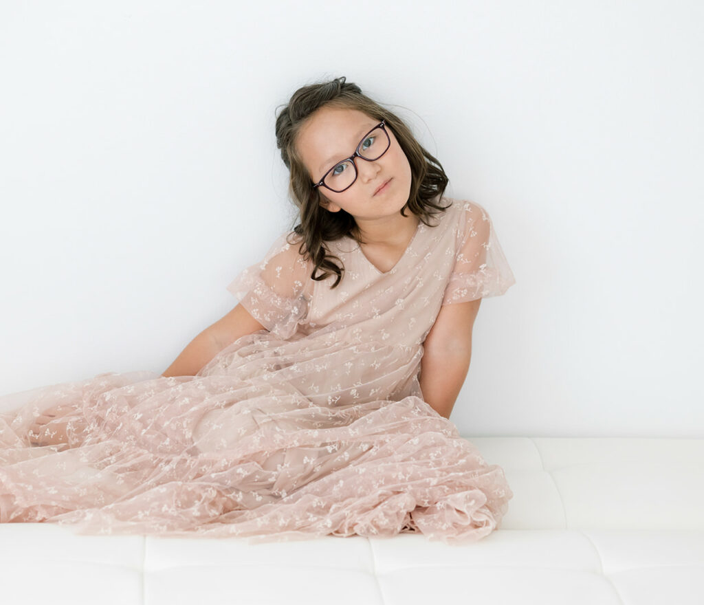 A young girl with glasses sits on a bed in a studio wearing a pink dress from Threadfare