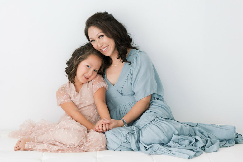 A mother in a blue dress sits on a bed in a studio holding hands with her toddler daughter in a pink dress