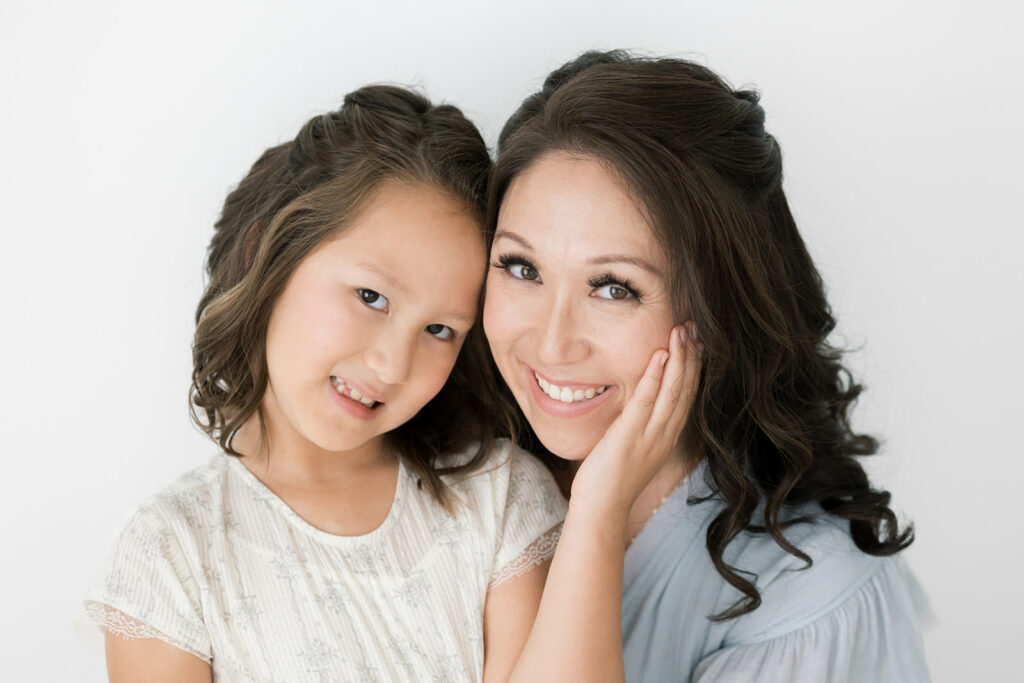 A happy mom sits in a studio touching heads and smiling with her toddler daughter