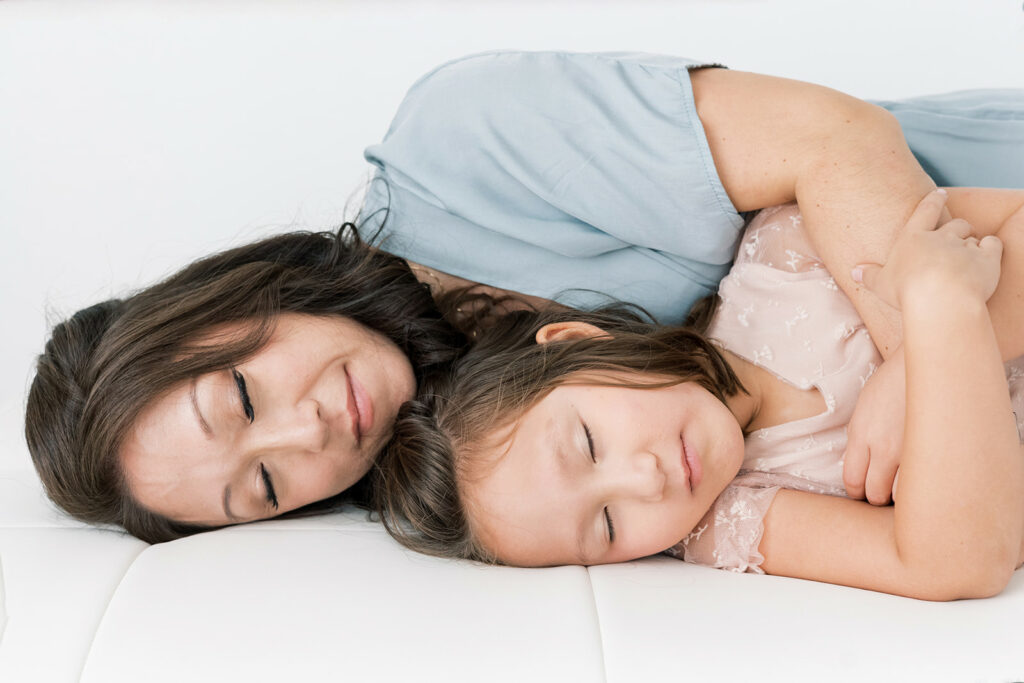 A happy mother cuddles with her toddler daughter while laying on a bed in a studio