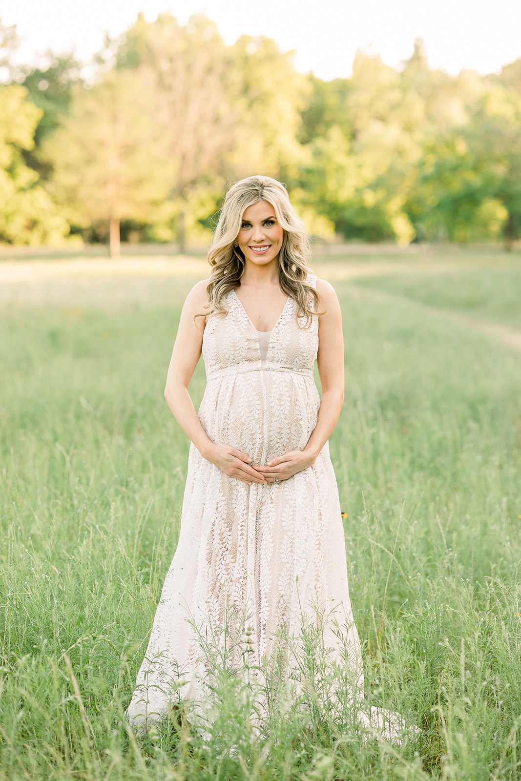 A mother to be in a lace maternity gown stands in a park field at sunset while holding her bump after meeting with Concierge Doulas of Houston