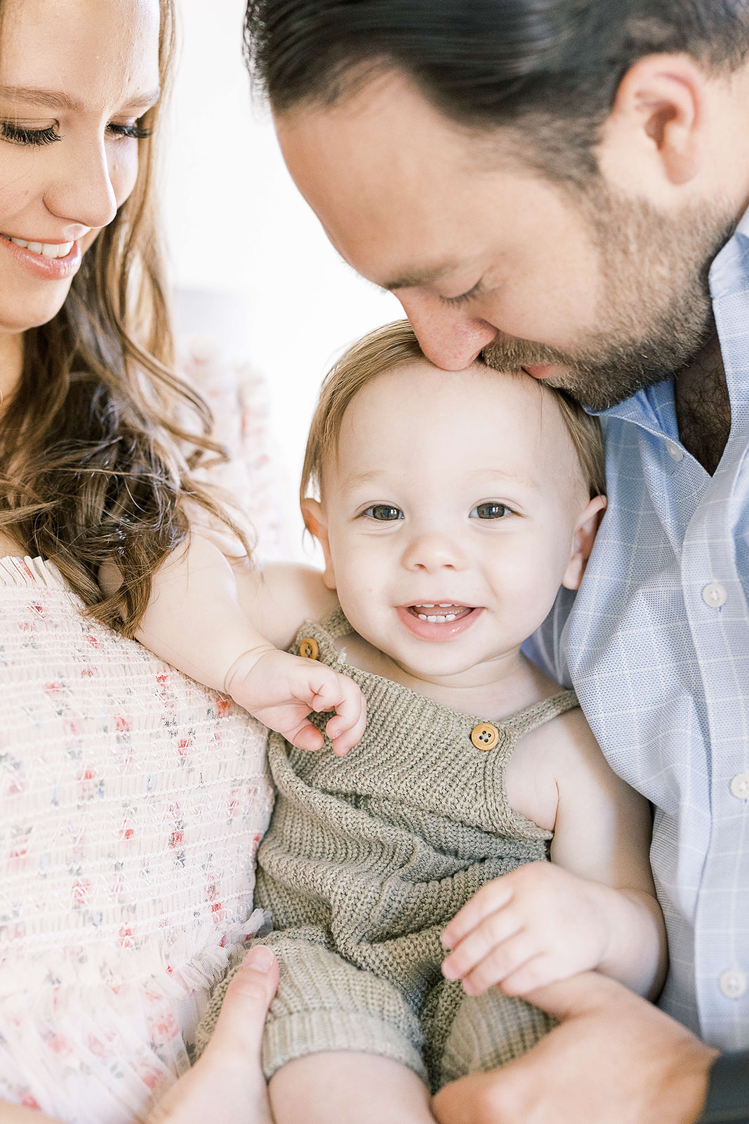 A father kisses the head of his toddler son smiling and sitting in mom and dad's arms