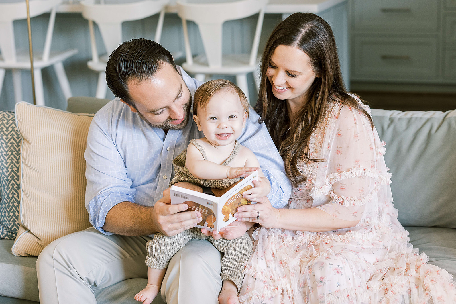 Happy mom and dad sit on a couch reading a bear book with their toddler son in dad's lap