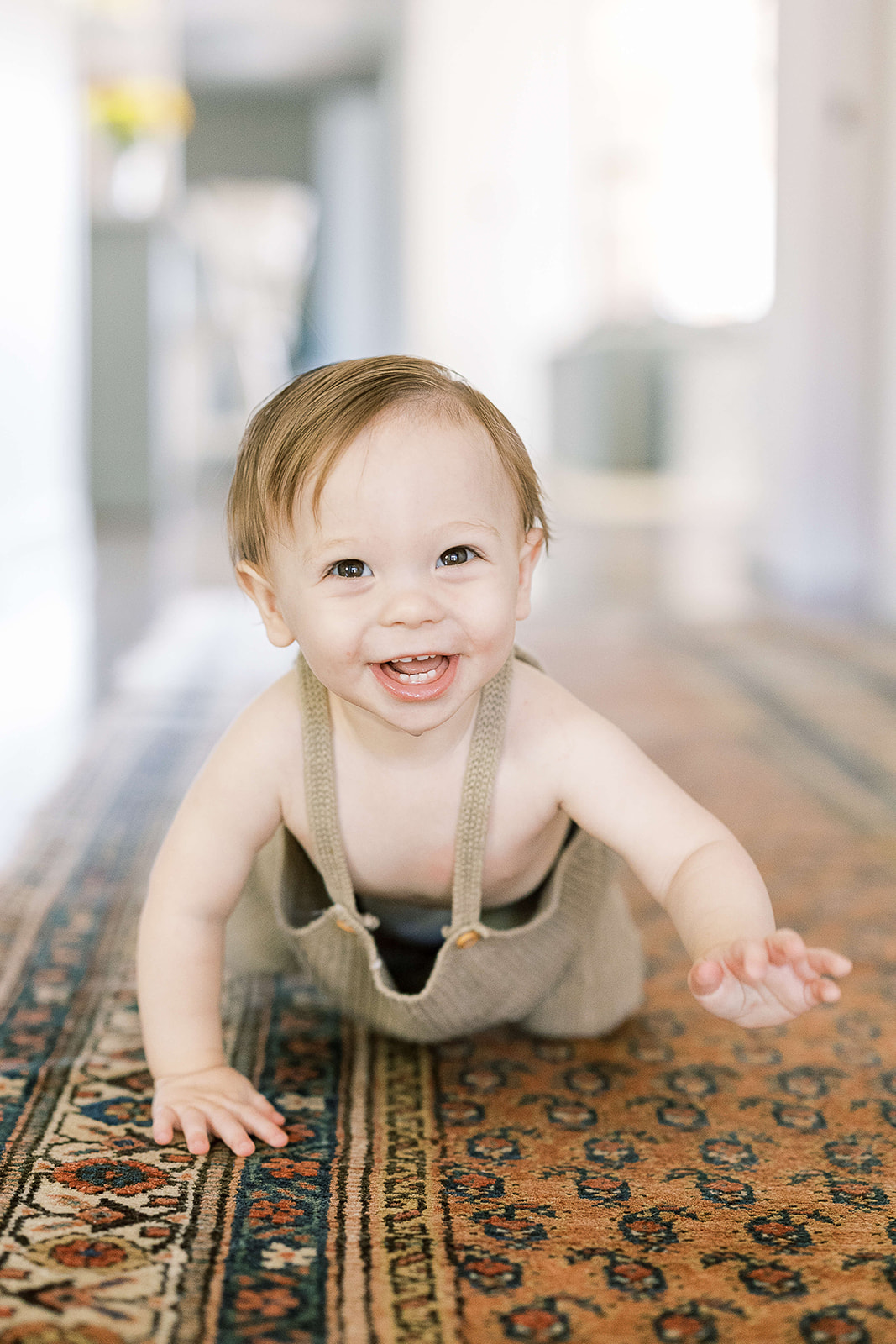 A toddler boy in brown overalls crawl down a hallway rug with a big smile after visiting Houston Baby Boutiques