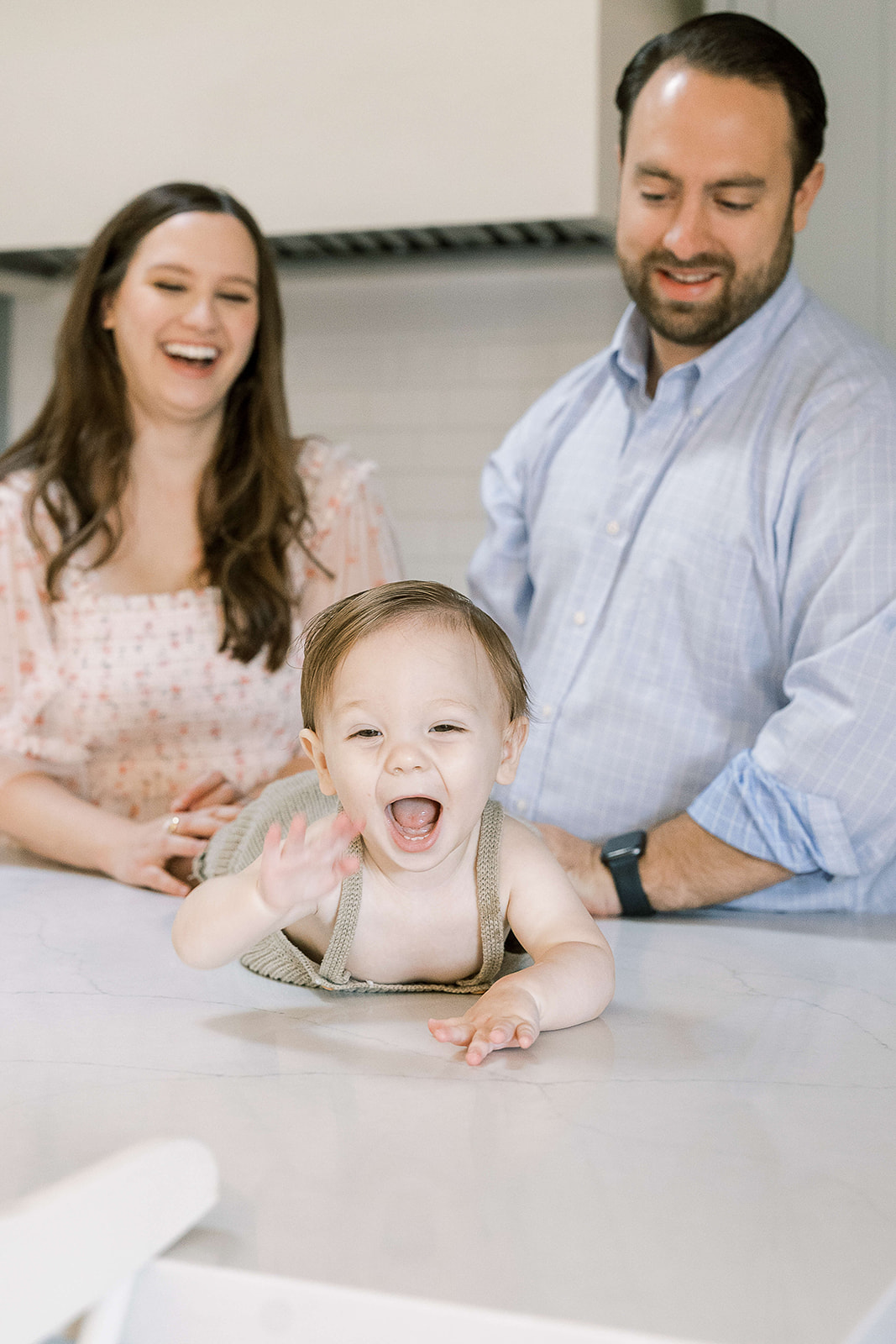 A happy toddler boy in green overalls crawls across a table with mom and dad laughing behind him after visiting Houston Baby Boutiques