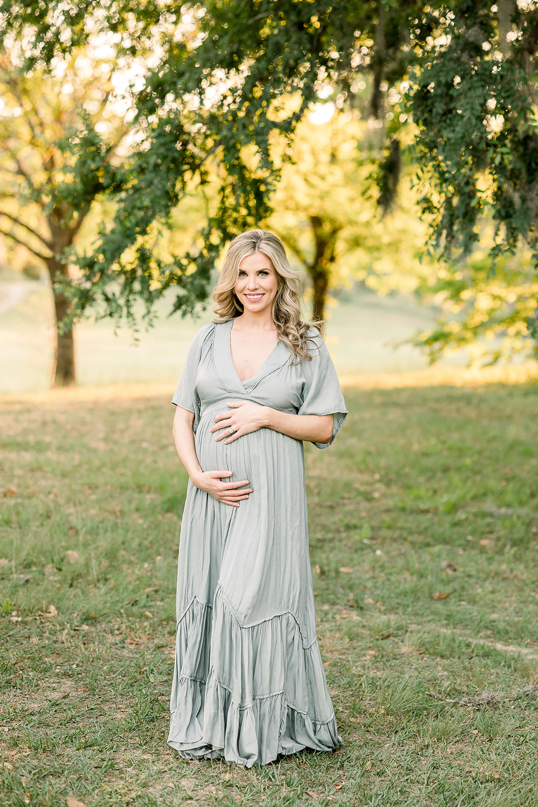 A smiling mom to be in a green flowing maternity gown stands under a tree in a park at sunset