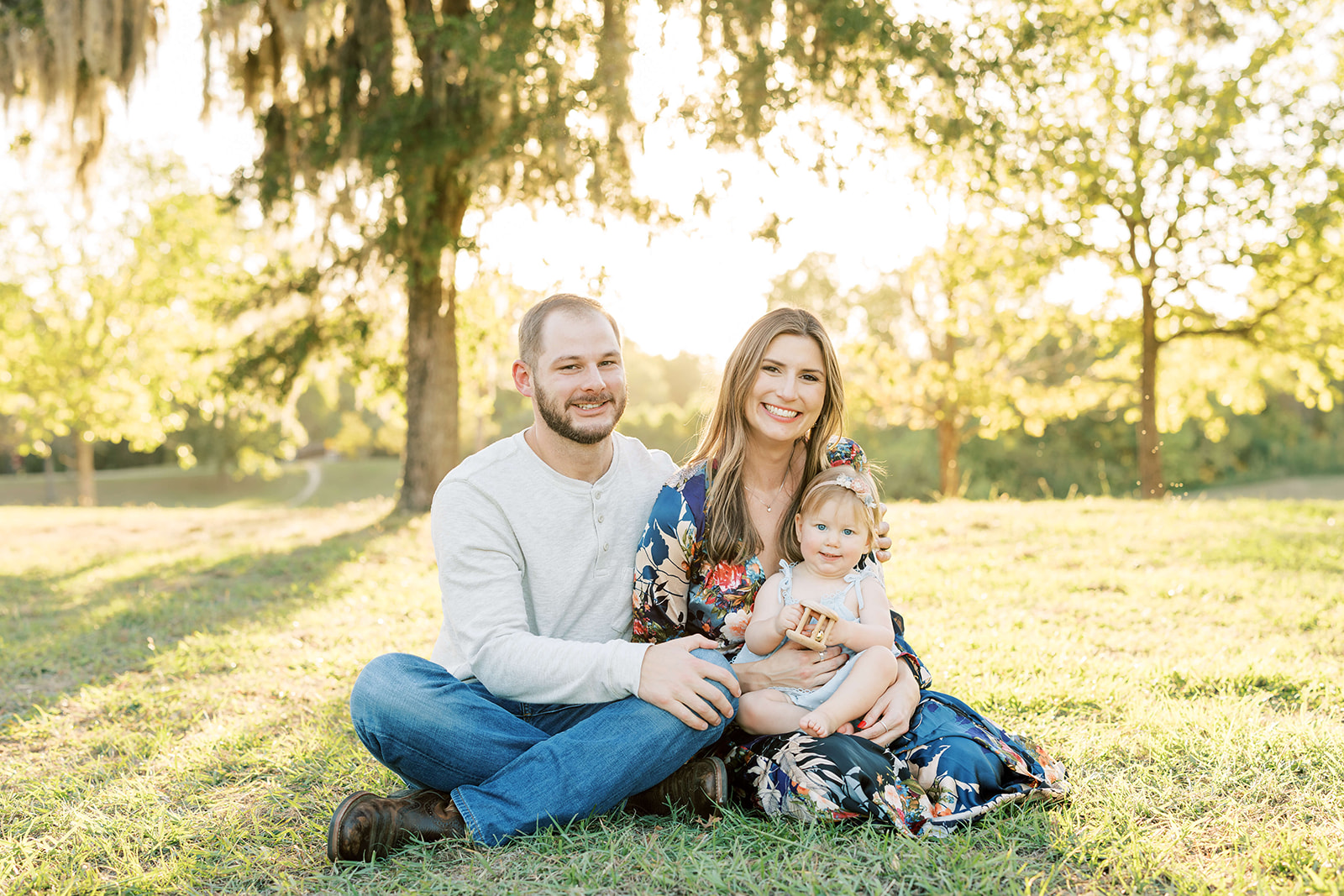 Happy parents sit in a park lawn playing with their toddler daughter and a wooden toy