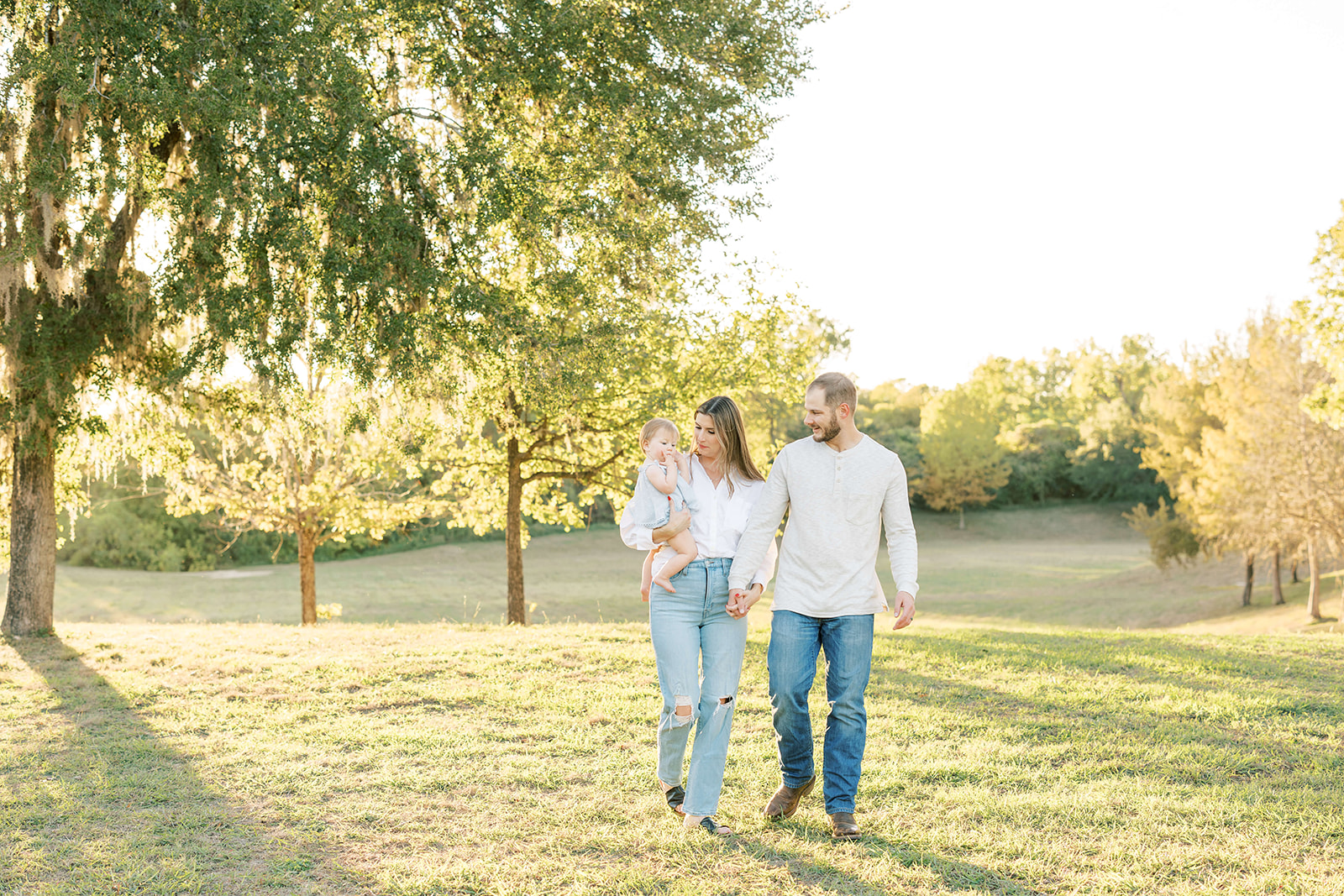 Mom and dad walk through a park lawn while playing with their toddler daughter in mom's arm