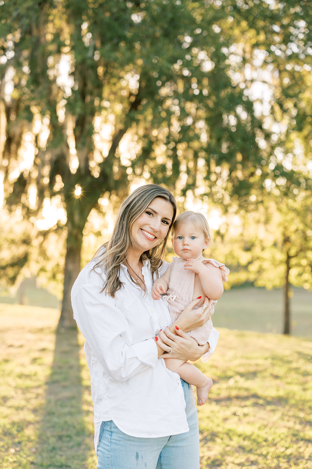 A happy mother stands under a tree at sunset while holding her toddler daughter on her hip before visiting Houston Daycares