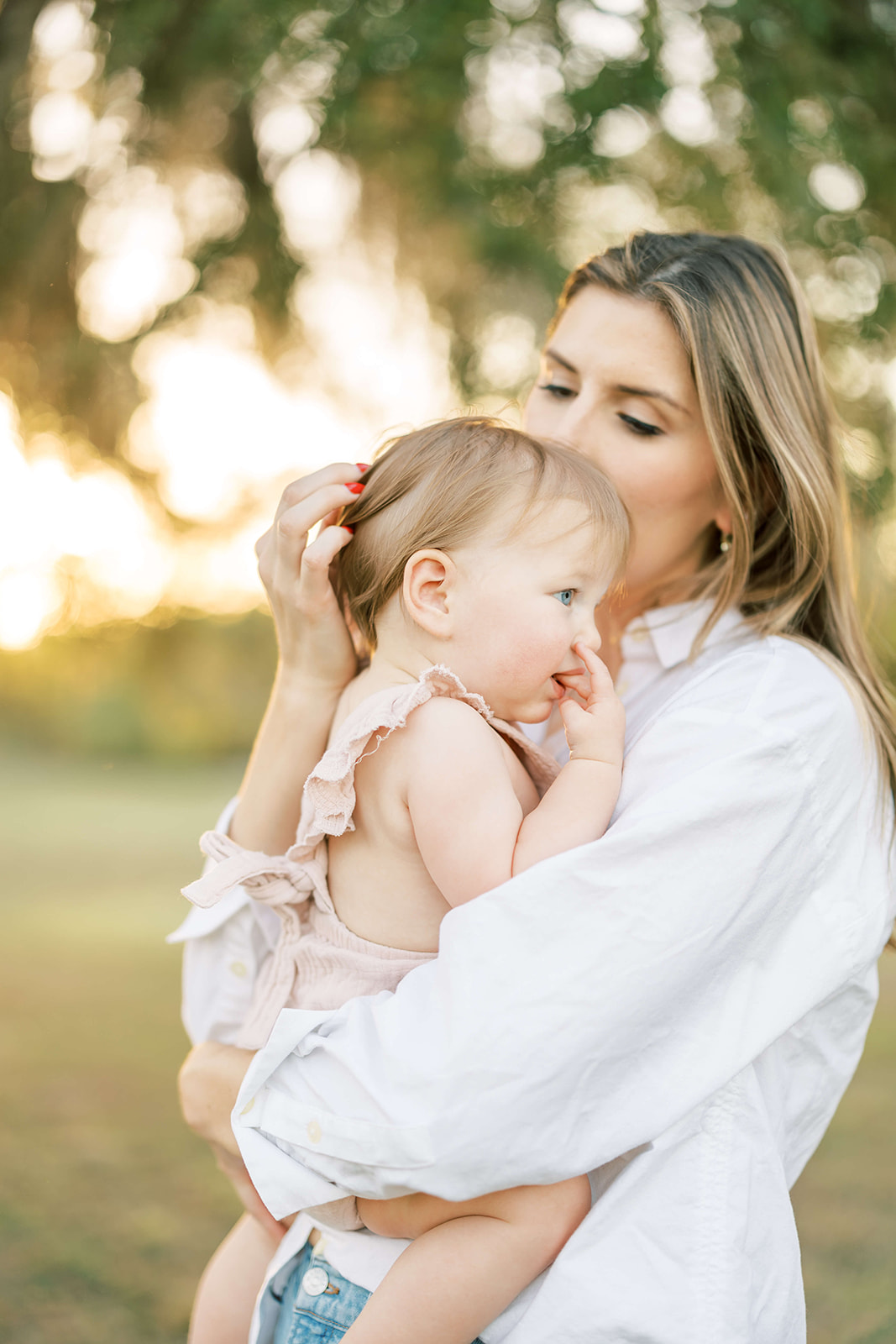 A mother in a white shirt holds her toddler daughter on her hip while exploring a park at sunset