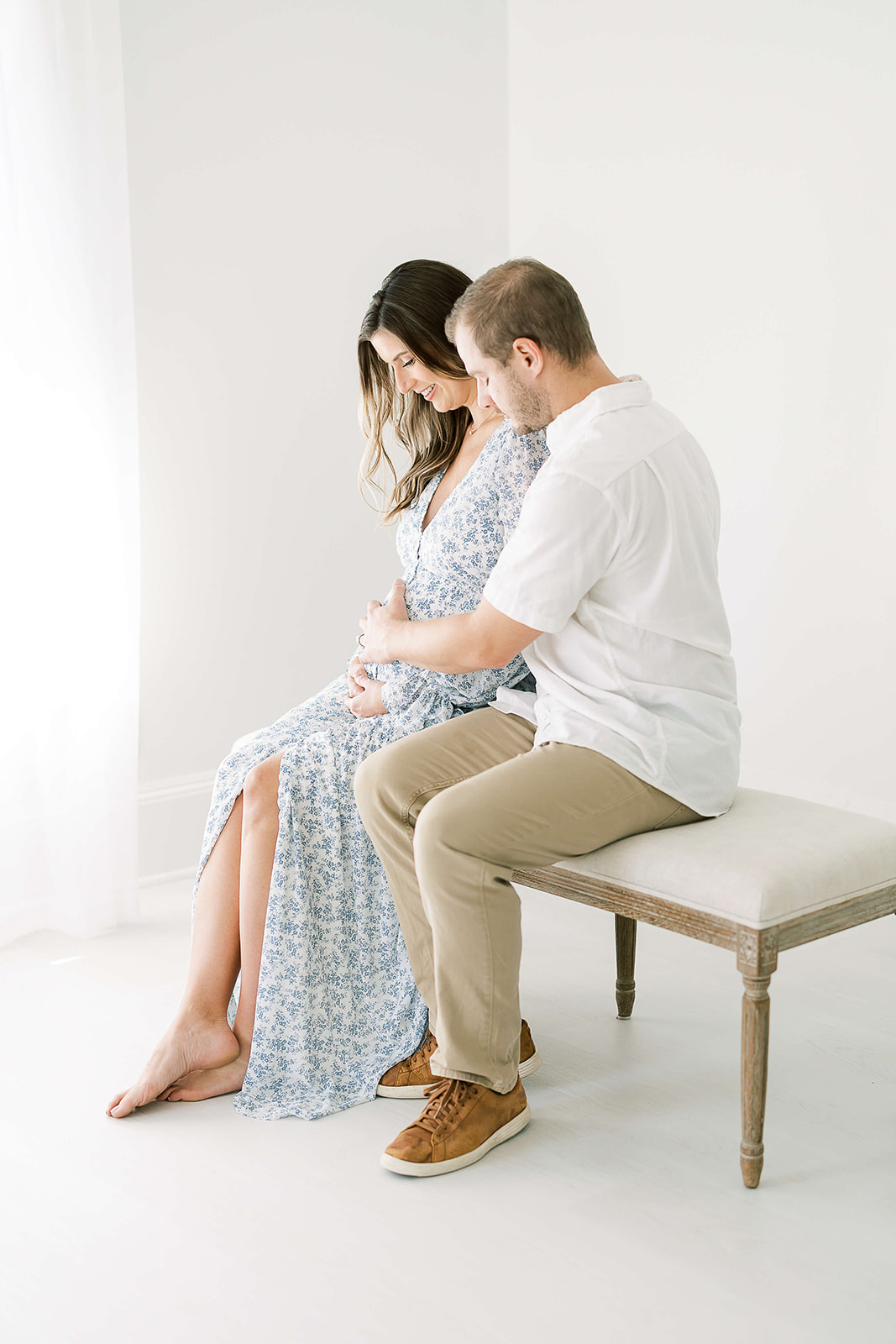 Expecting parents sit on a bench in a studio holding and smiling down at the bump