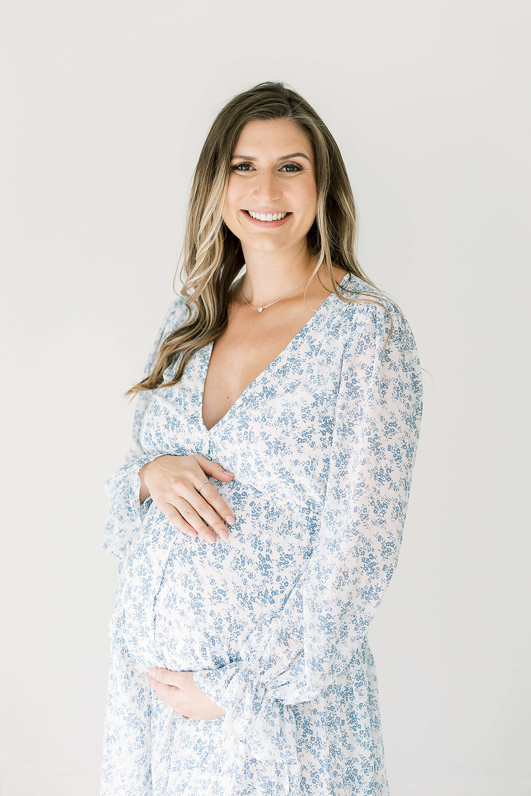 A mother to be in a blue floral print maternity dress smiles big while standing in a a studio holding her bump after visiting Houston OBGYN