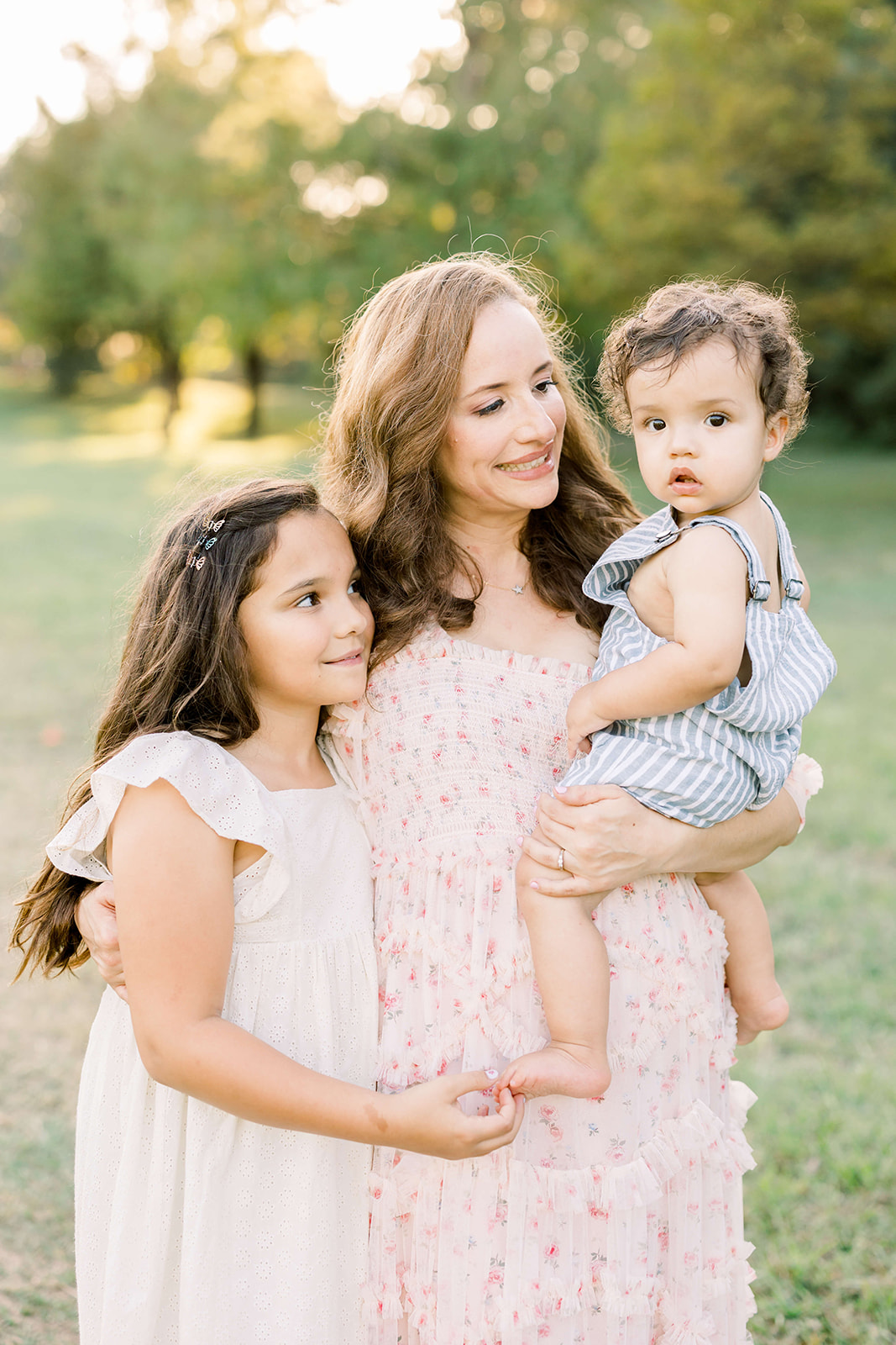 A happy mom smiles big while standing in a park with her toddler daughter hugging her and younger son on her hip in a pink dress