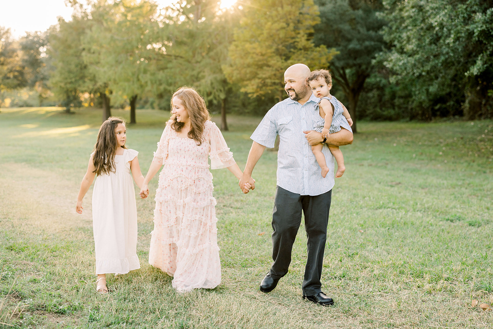 Happy mom and dad walk through a park lawn at sunset holding hands and holding their toddler son before visiting Houston pediatric dentists